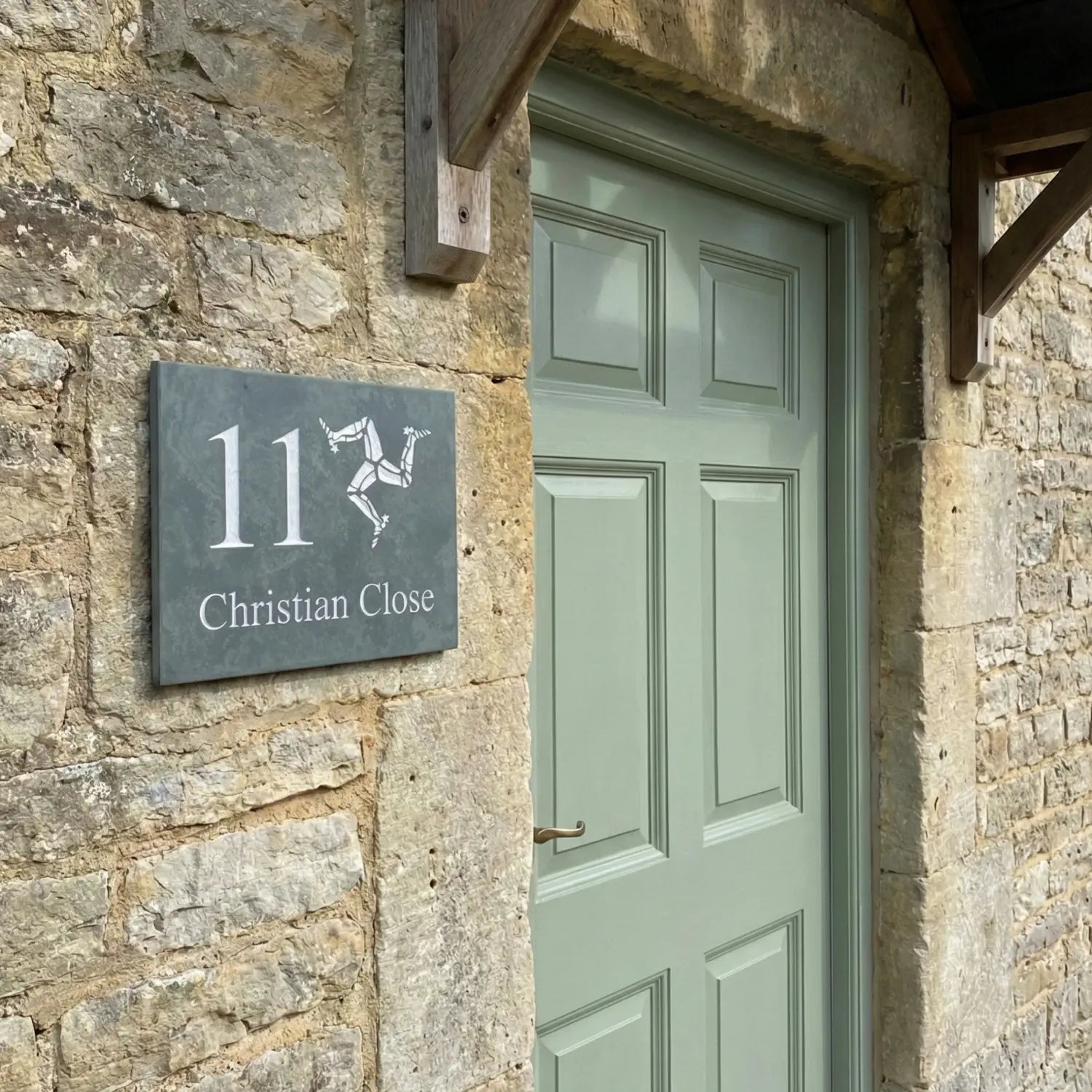 House number sign with Isle of Man triskelion on a stone wall next to a window with a reflection of a garden