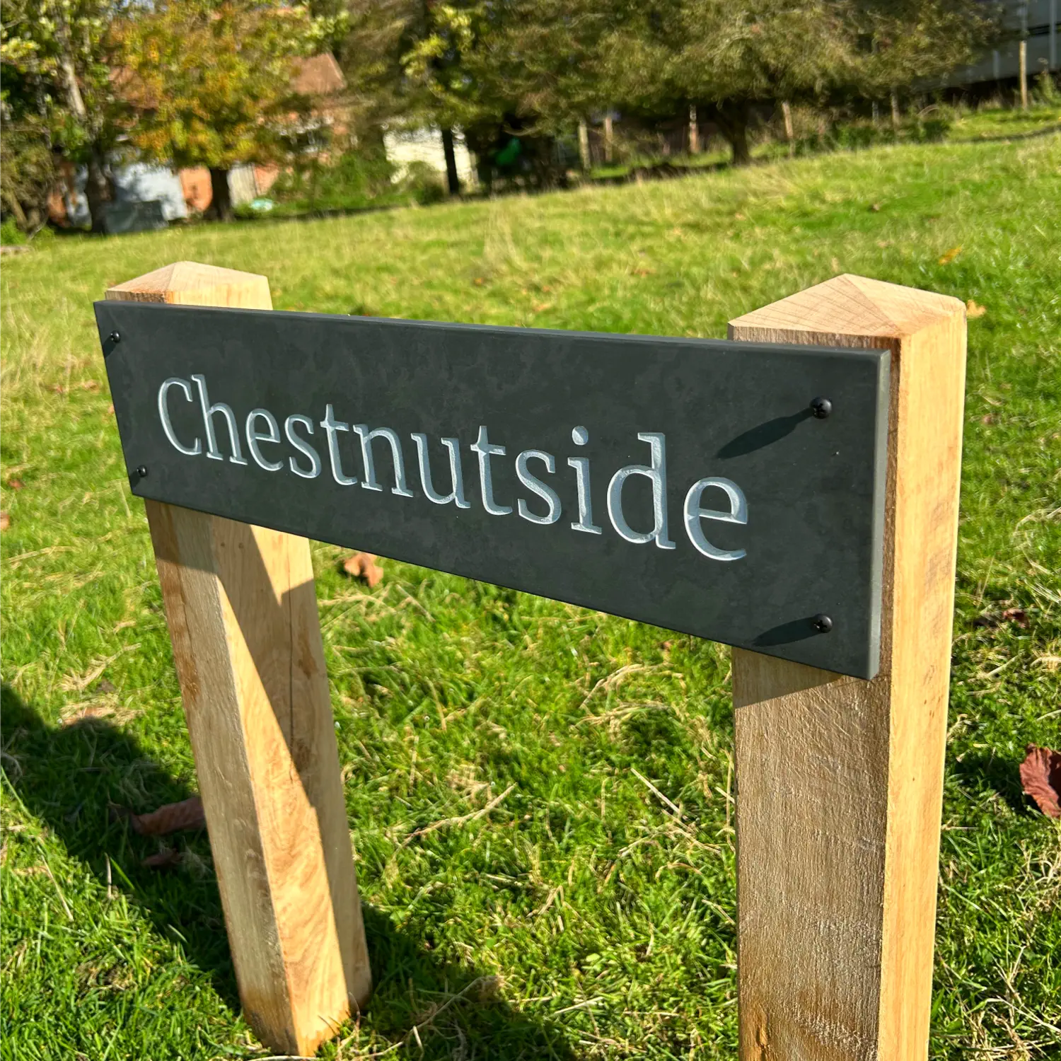 The image shows a slate house sign mounted onto sturdy oak posts with pyramid tops. The sign reads “Chestnutside” in clear white lettering, and the oak posts are set into a grassy field. The overall appearance is stylish and rustic, showcasing the natural wood and elegant slate combination. The Bespoke Sign House logo is visible in the top right corner.