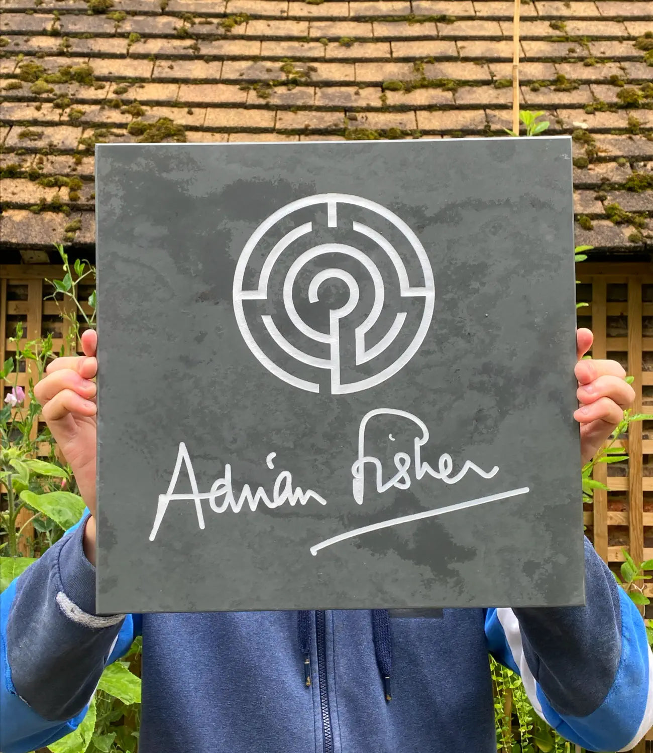 A custom slate sign featuring an engraved white maze design and the signature “Adrian Fisher,” held up outdoors against a rustic wooden trellis and mossy tiled roof background.