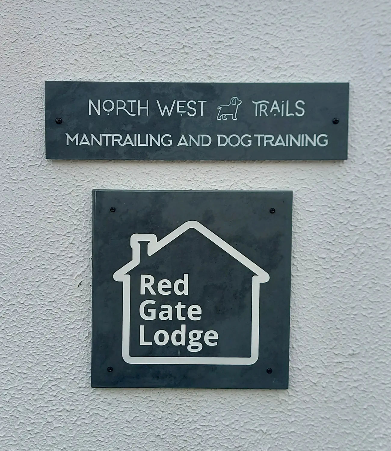 Two slate business signs mounted on a textured white wall. The top sign reads “North West Trails - Mantrailing and Dog Training” with a small dog icon, while the bottom sign features a house-shaped logo with “Red Gate Lodge” engraved inside.