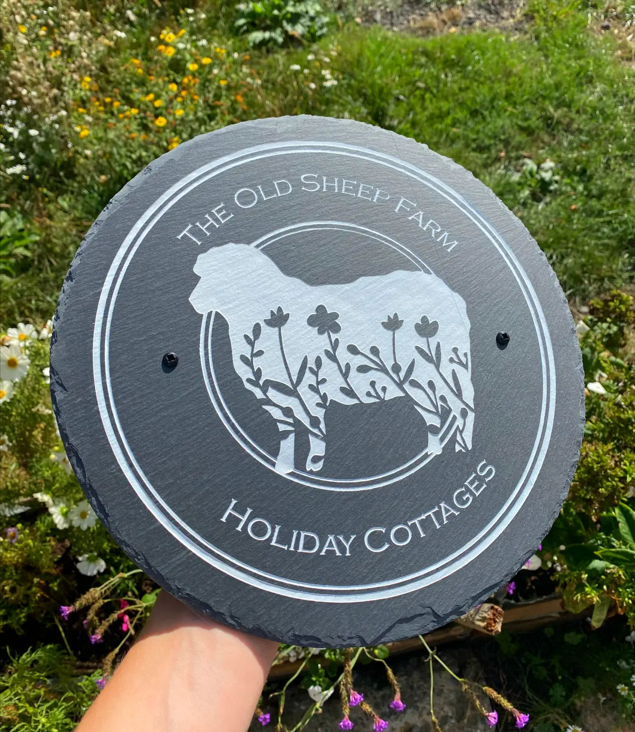 A custom round slate sign featuring an engraved sheep design with floral detailing, along with the text “The Old Sheep Farm Holiday Cottages,” held outdoors against a backdrop of wildflowers and greenery.