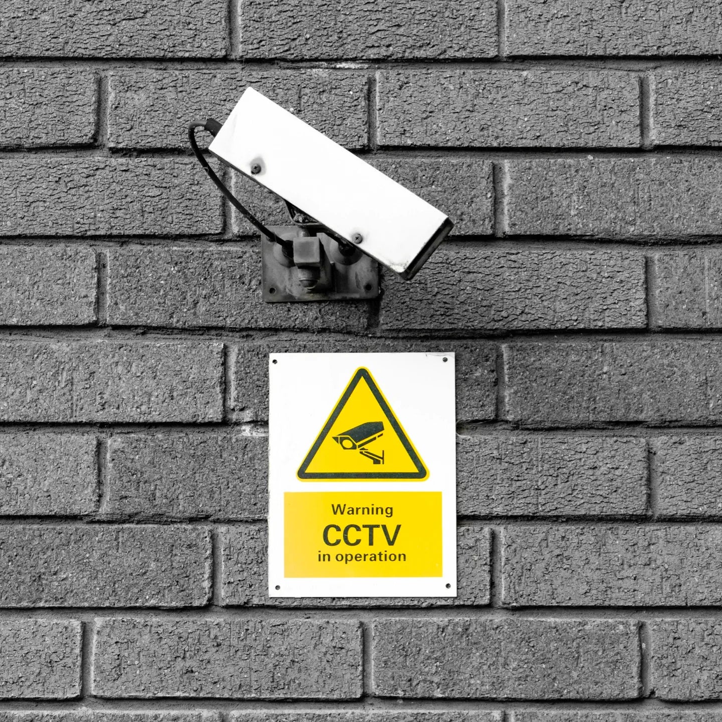 A CCTV camera mounted on a gray brick wall above a yellow warning sign that reads, “Warning: CCTV in operation.”
