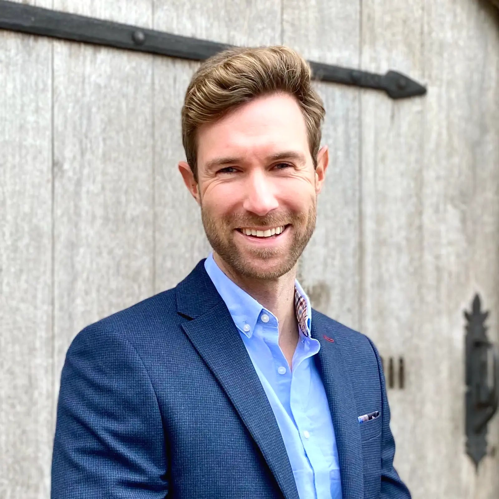 Man in a blue blazer and light blue shirt standing against a wooden background