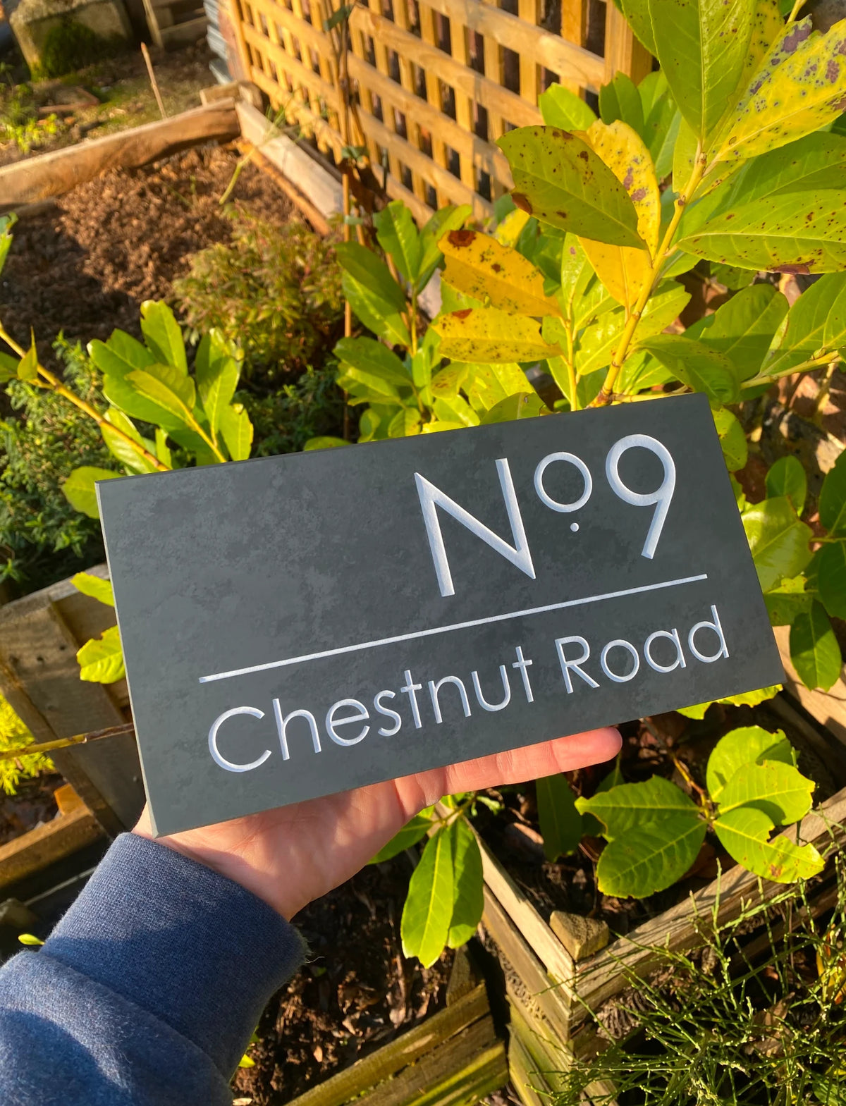 A dark slate-style house number sign reading 'No 9 Chestnut Road' held by a person's hand in a sunny garden with green plants and wooden raised beds in the background.