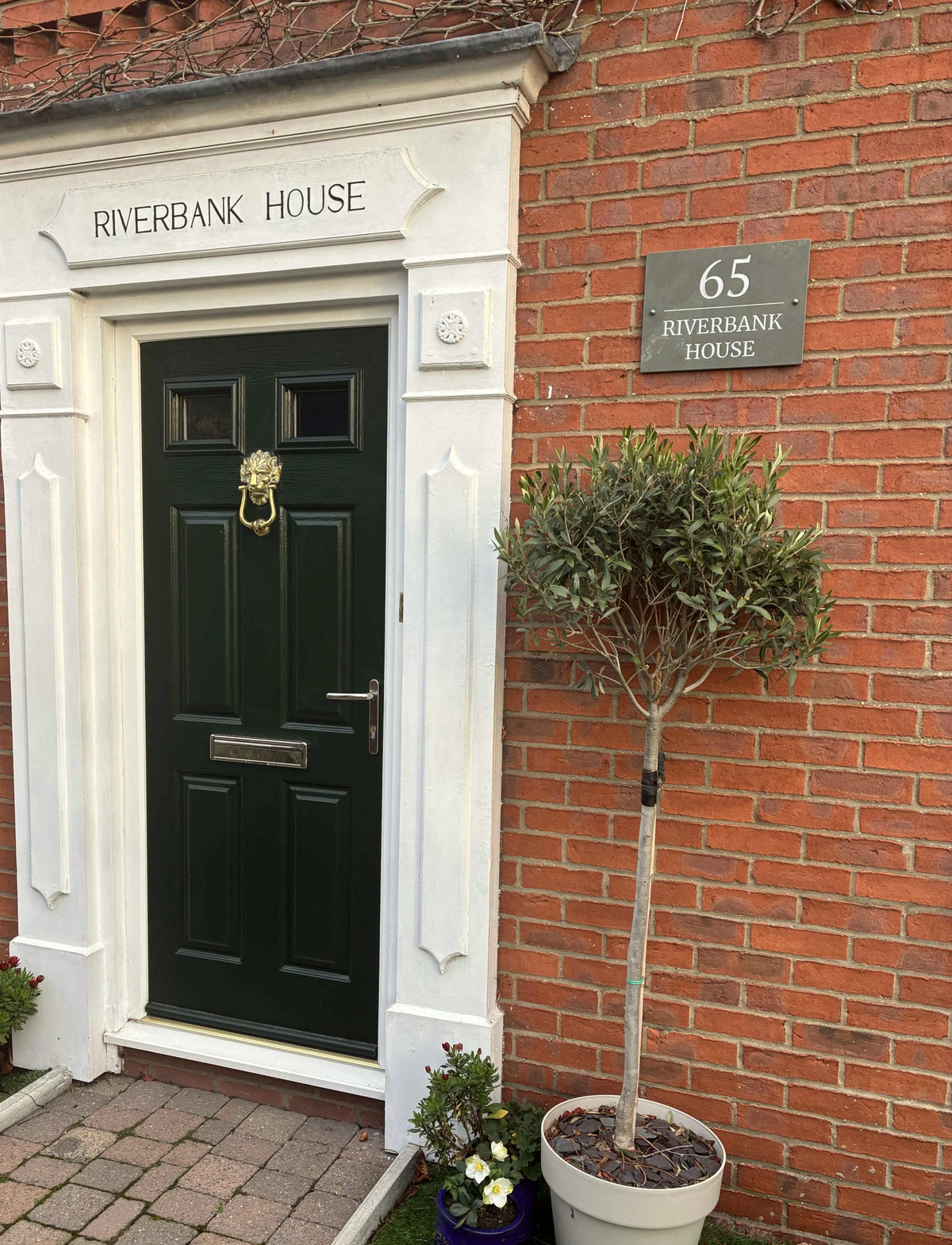 A dark green wooden front door with a brass lion knocker, set in a white stone frame on a brick house. A brick address plaque reads '65 RIVERBANK HOUSE', and a potted topiary tree stands to the right.