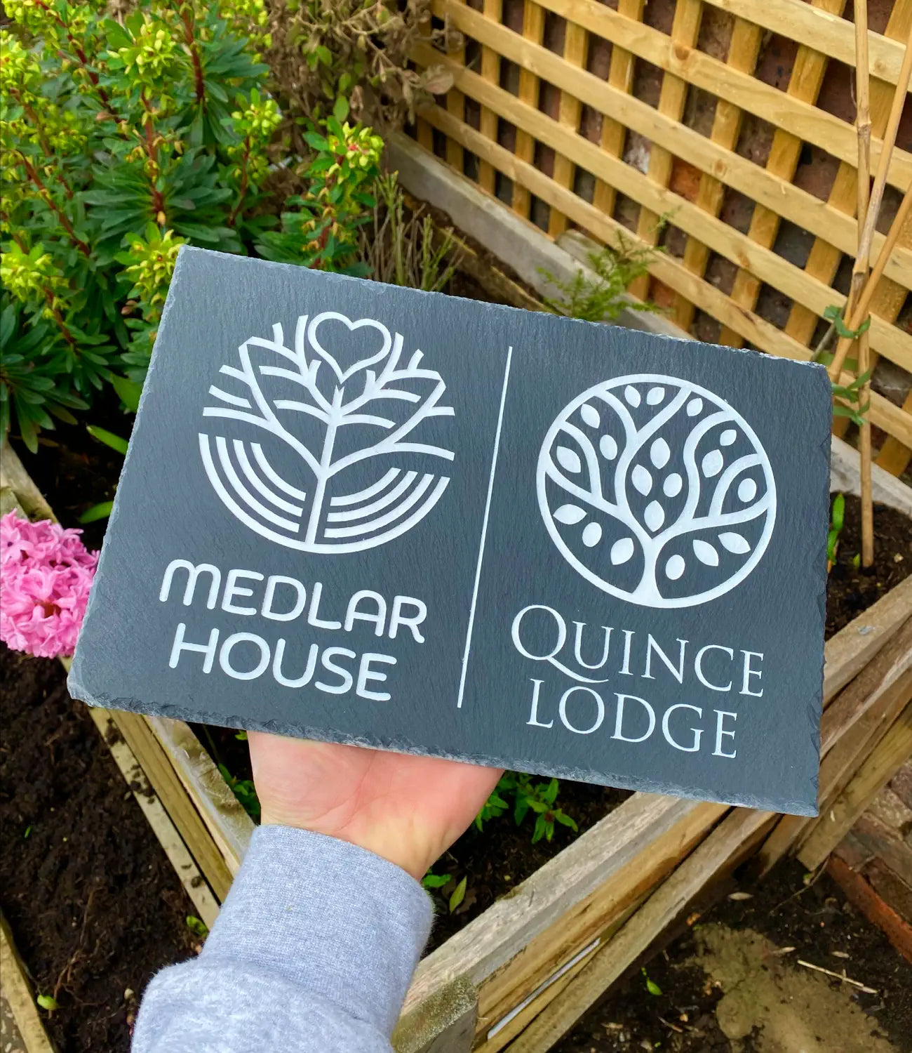 A custom slate house sign featuring engraved white botanical designs for “Medlar House” and “Quince Lodge,” being held outdoors against a wooden trellis and garden background.