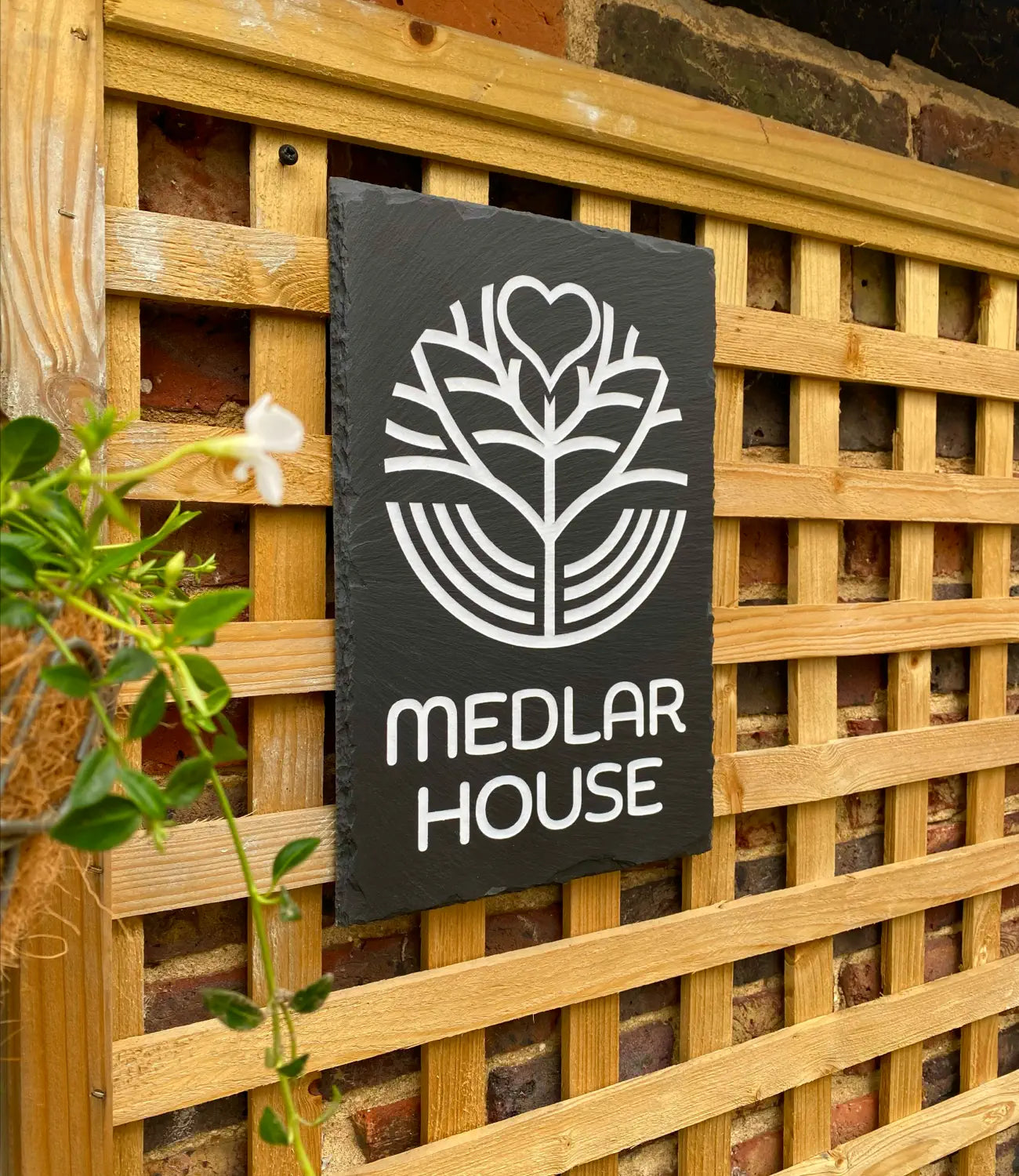 A custom slate house sign reading “Medlar House,” featuring an engraved white botanical design, mounted on a wooden lattice fence against a brick wall with a potted plant nearby.
