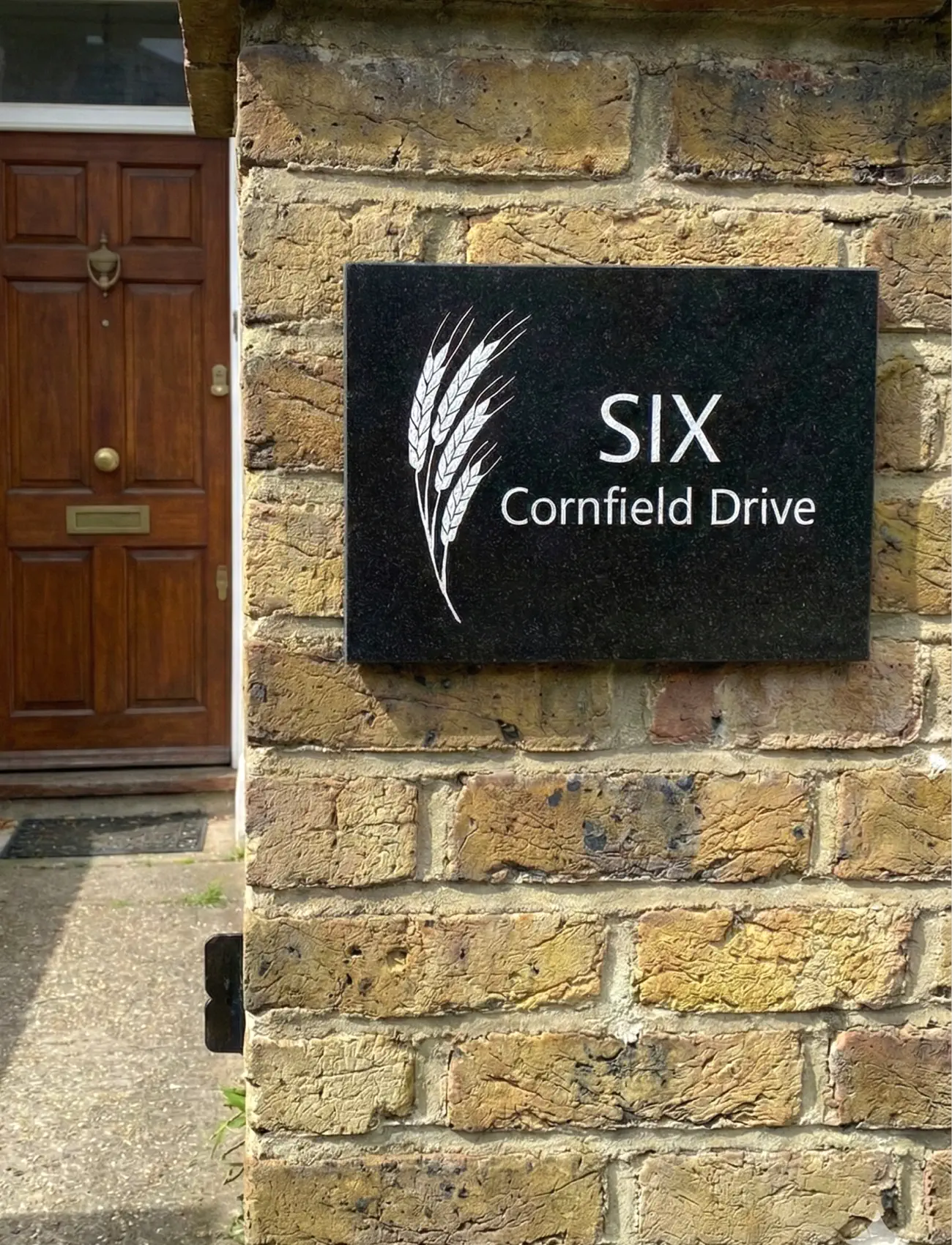 House number sign 'SIX Cornfield Drive' on a brick wall next to a wooden door.