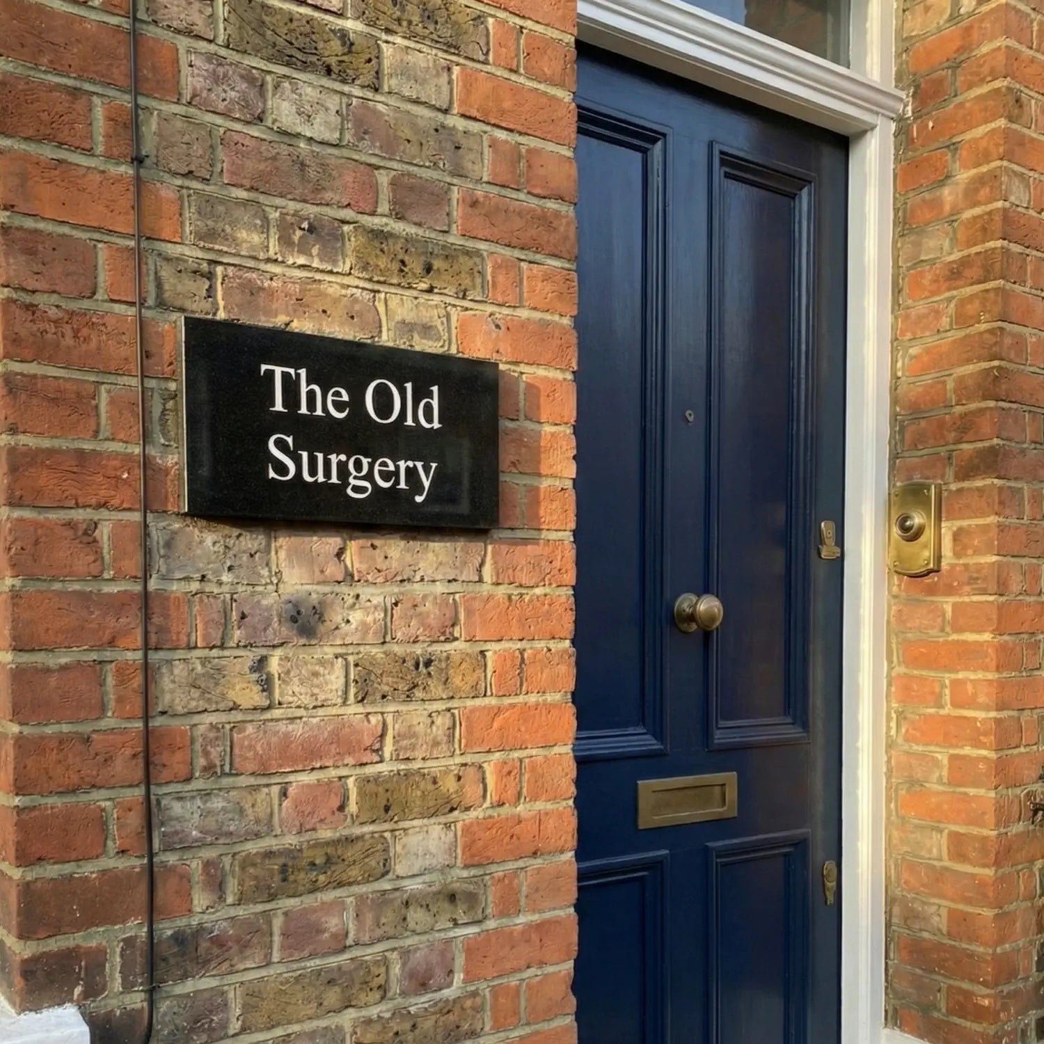 Black granite sign with 'The Old Surgery' on a brick wall next to a door.