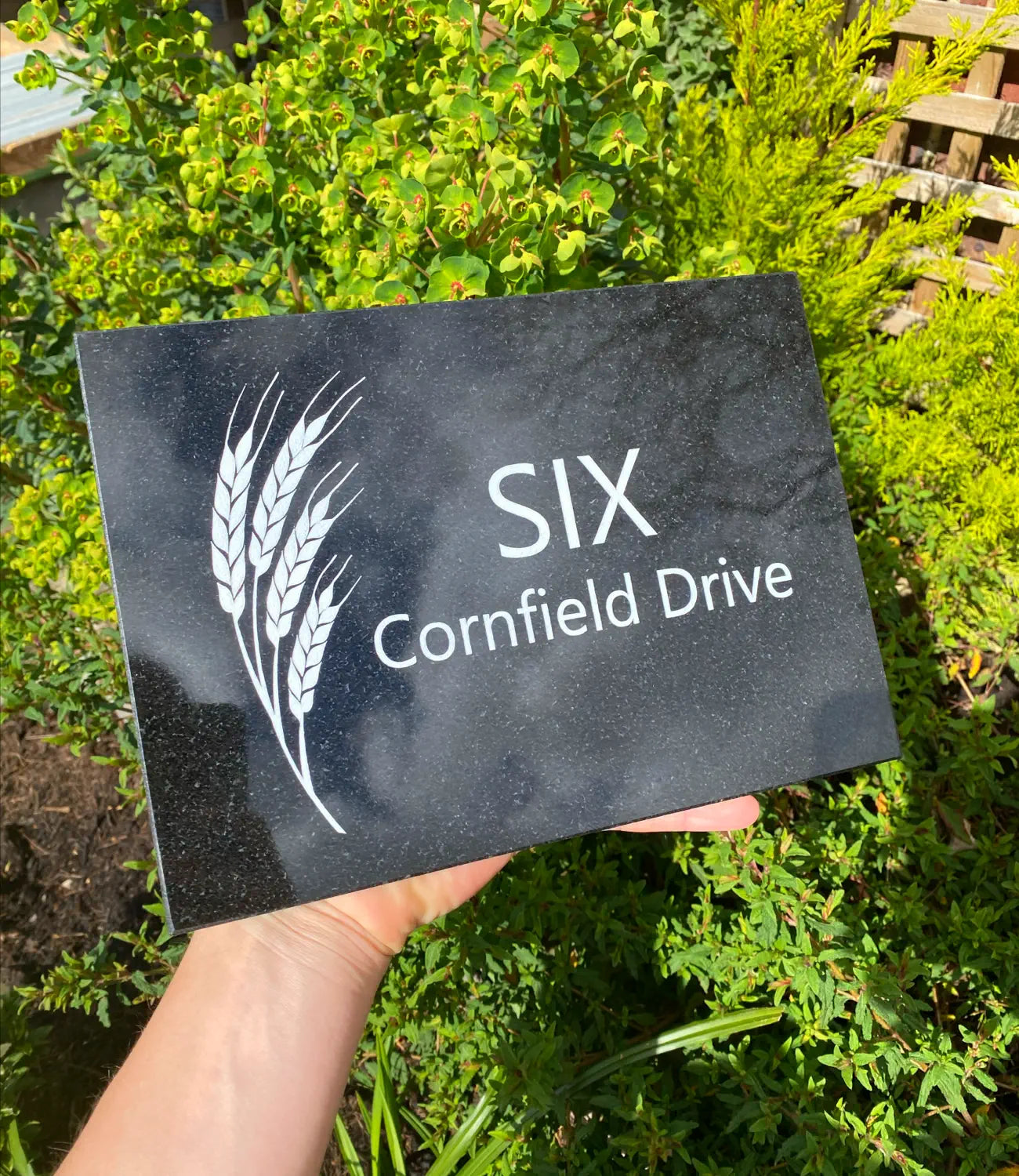 A custom black granite house sign with engraved white text reading “SIX Cornfield Drive,” featuring a wheat design, held outdoors against a lush green garden background.