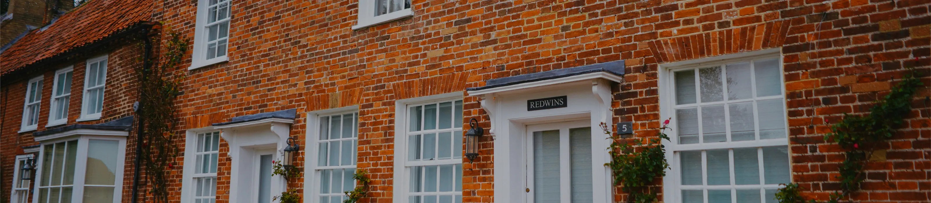 A banner featuring a variety of house signs, including wall-mounted and freestanding designs, crafted in slate with elegant text. The signs are displayed on different backdrops, such as brick, wood, stone, and painted walls. A man is shown smiling and pointing at one of the signs.