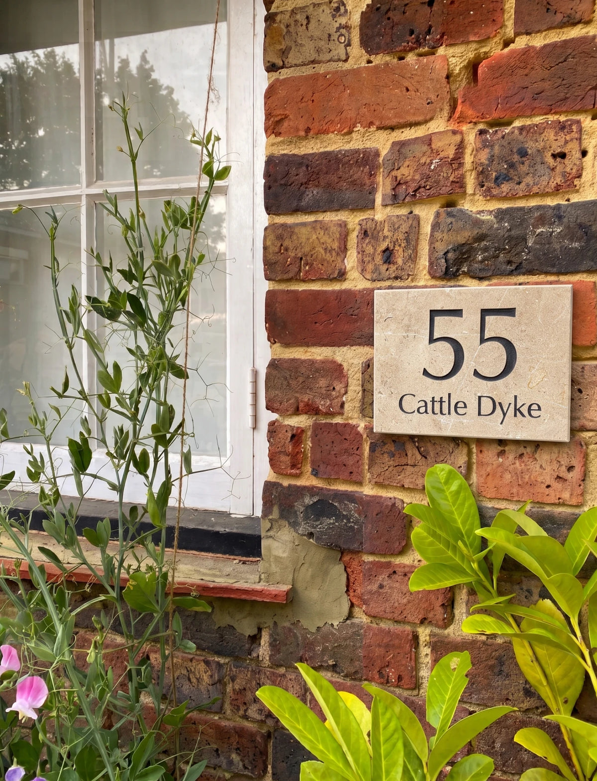 A square beige house number plaque reading '55 Cattle Dyke' mounted on a red and brown brick wall, with green plants in the foreground and a white window frame to the left.