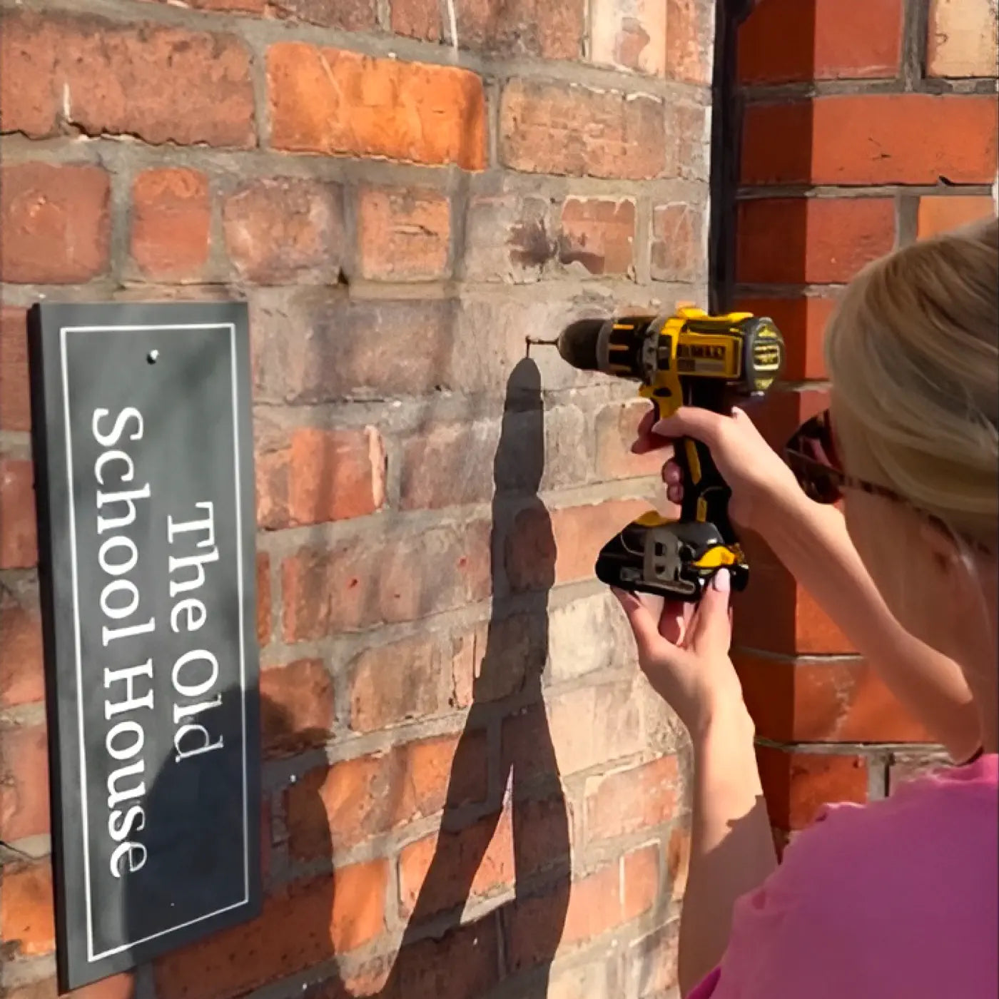 Person drilling into a brick wall to install a house sign that reads “The Old School House