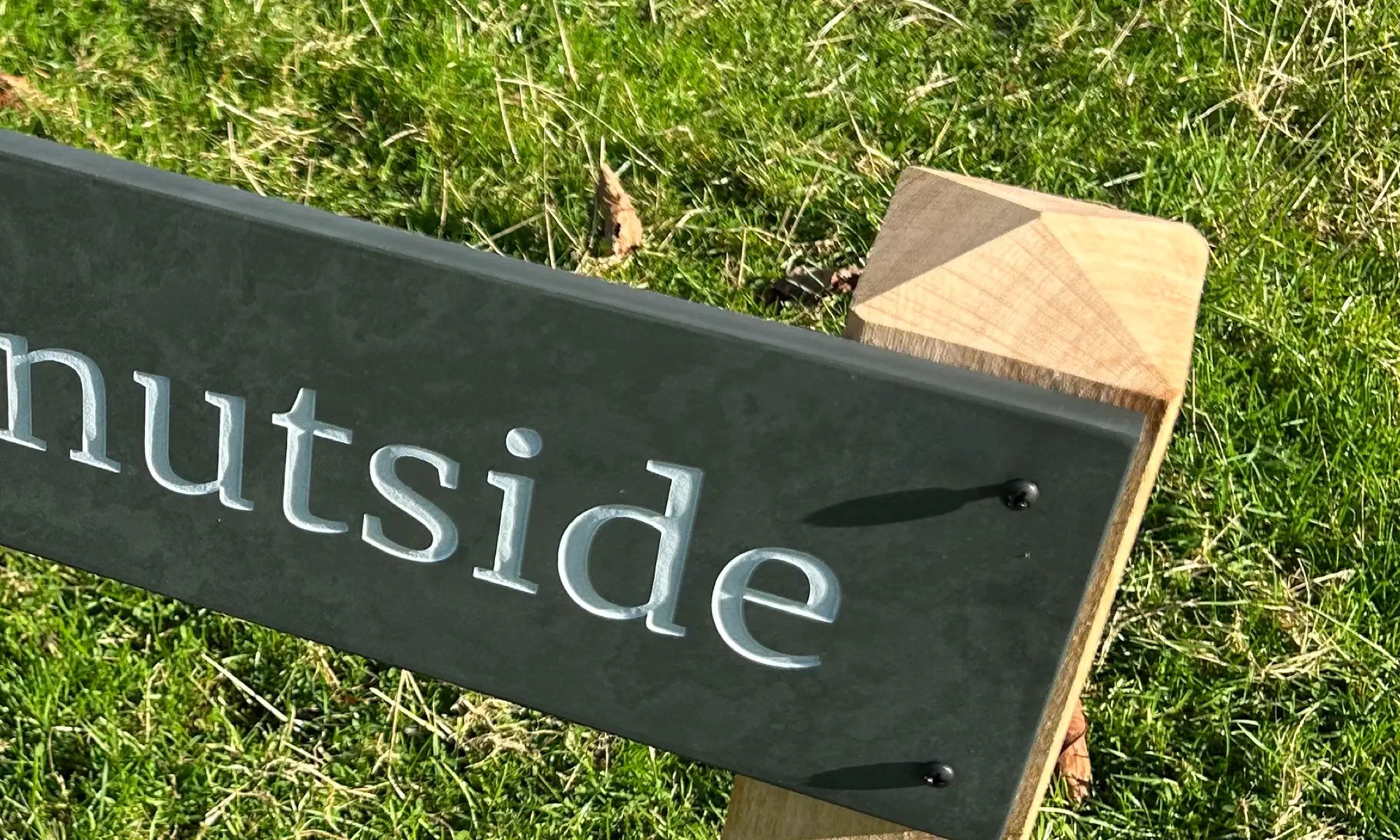 Close-up of a slate house sign with engraved lettering, mounted on an oak post with a pyramid top, set against a grassy background.