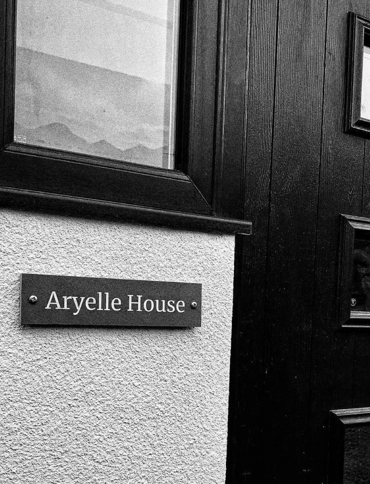 Black and white photograph of a house exterior showing a dark wooden door with small windows, a textured light wall, and a sign reading 'Aryelle House'.