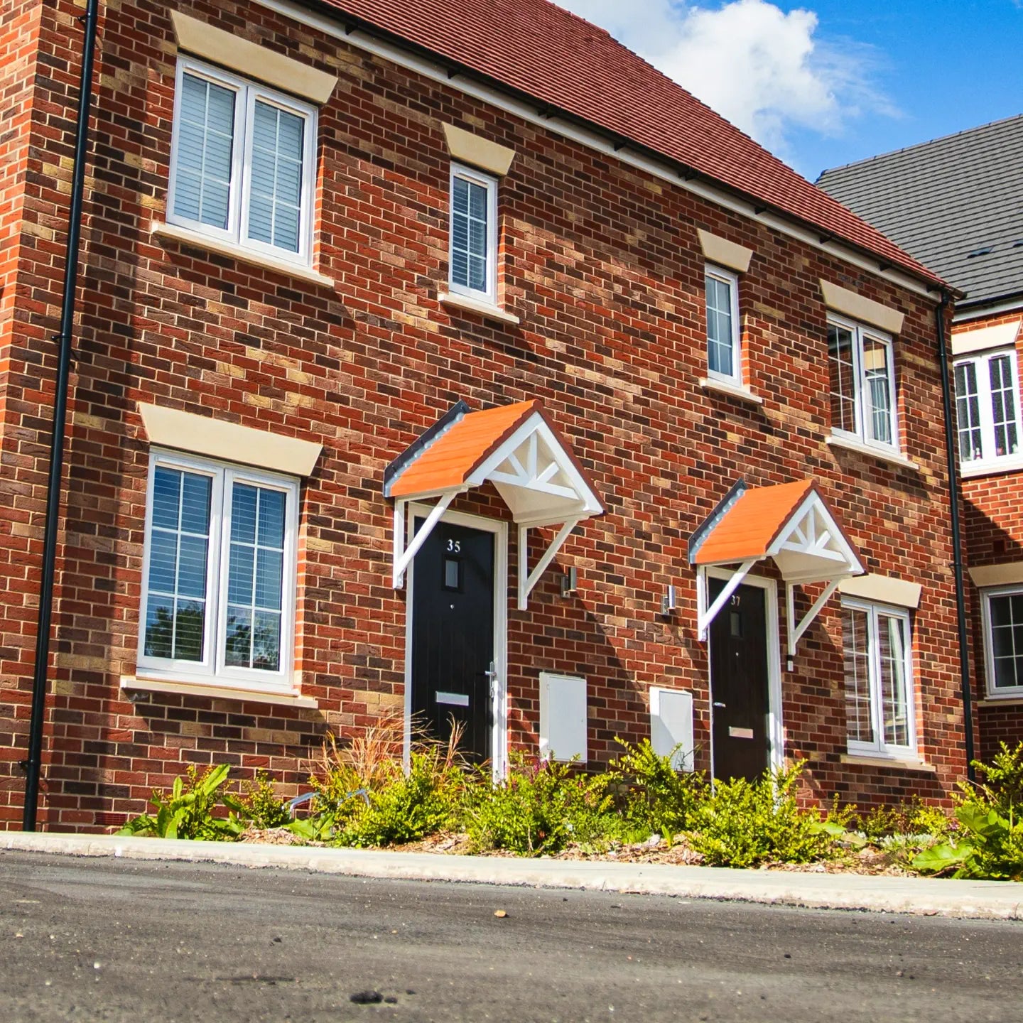A pair of modern red-brick townhouses with white-framed windows and black front doors, each featuring small triangular wooden porch coverings with orange roofs. A neatly landscaped front garden with green shrubs and plants lines the pathway. The scene is bright and sunny with a clear blue sky in the background.
