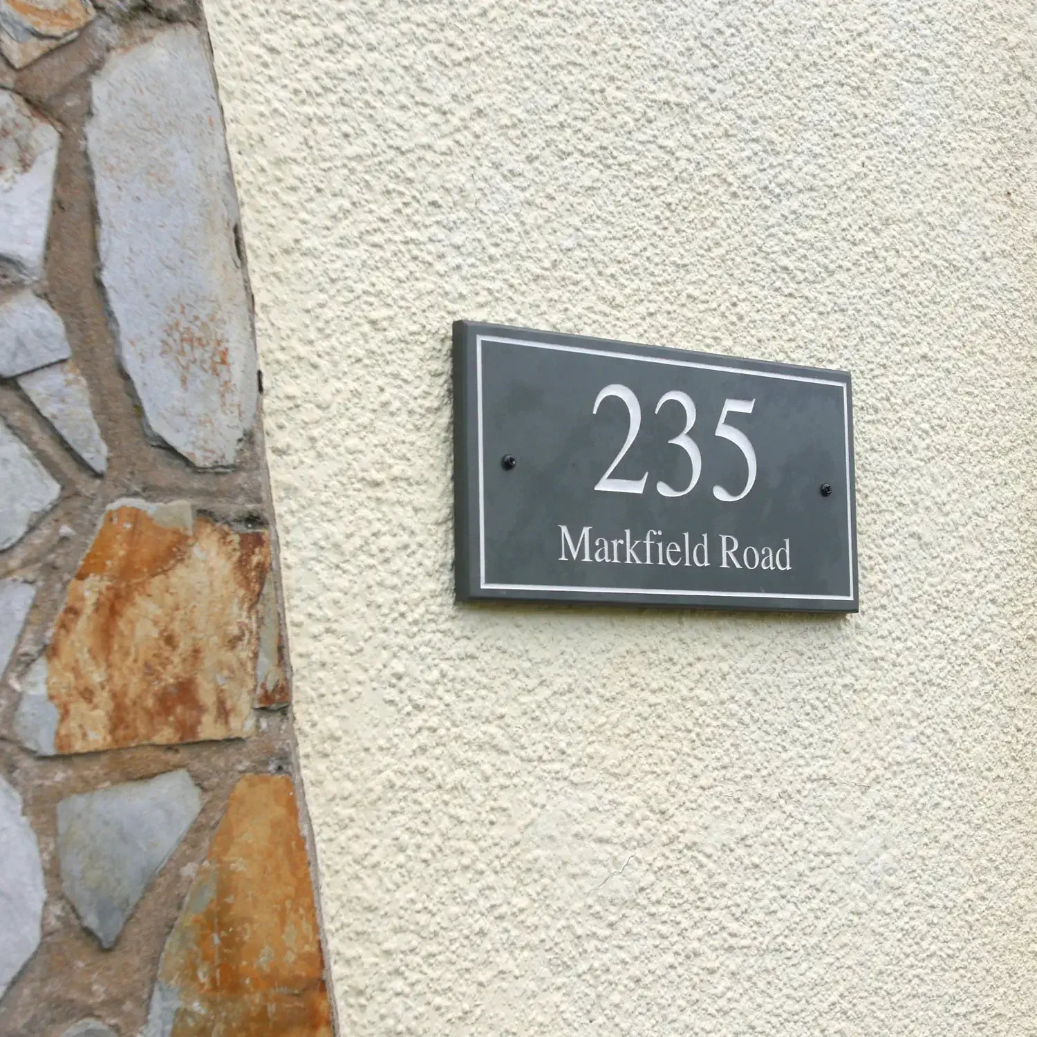 a slate house sign with a border design on a beige pebble-dashed UK house. The sign has engraved numbers and letters that are painted white