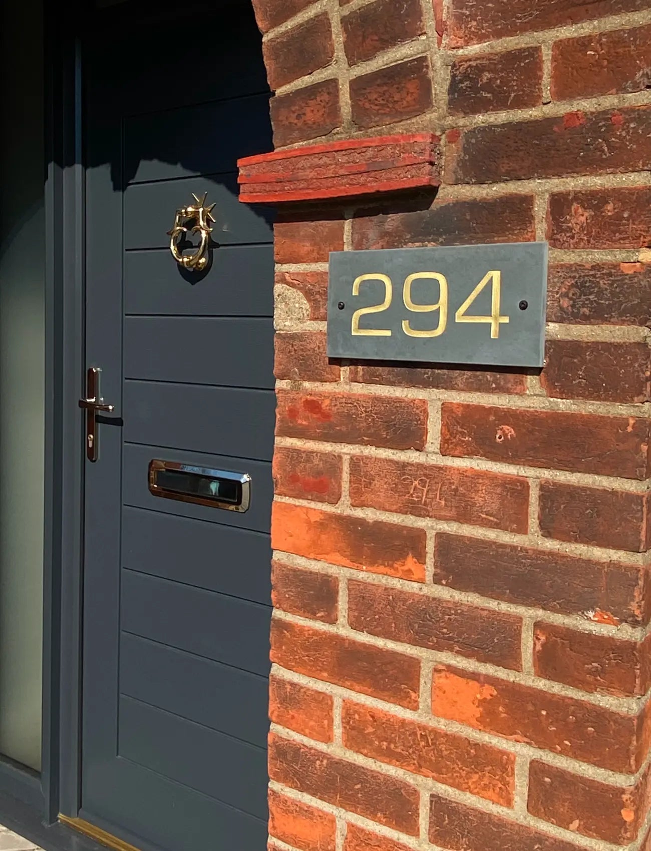 Dark gray door with gold handle and address number 294 on a brick wall.