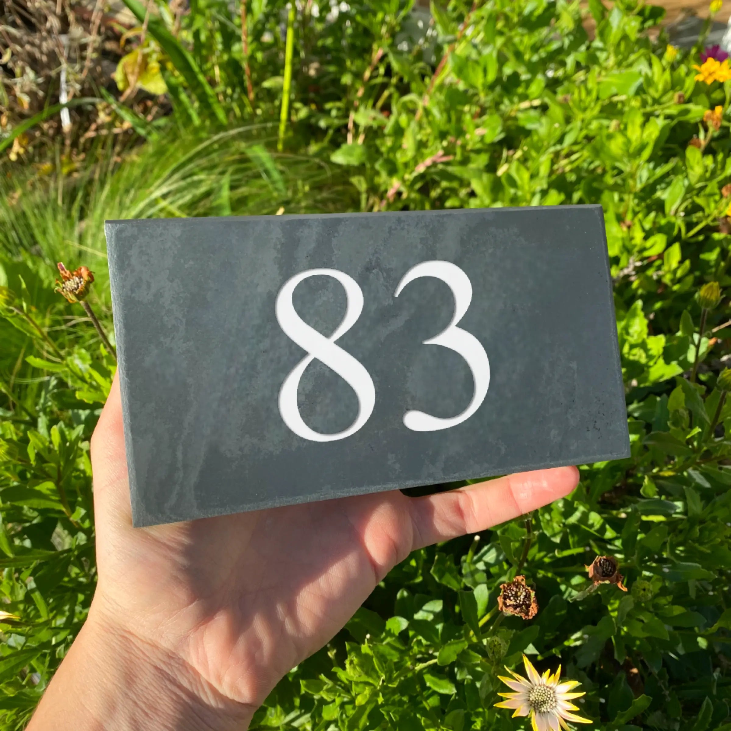 This image shows a hand holding a rectangular slate sign with the number "257" engraved in large white font. The background features a wooden lattice fence with bricks visible behind it.