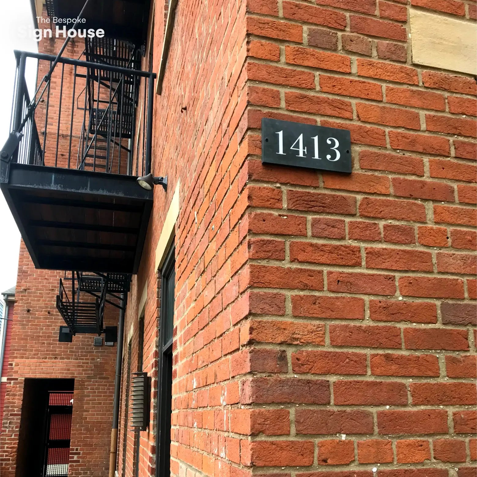 This image shows a rectangular slate sign with the number "1413" in white font mounted on a red brick wall at the corner of a building. The building features black metal fire escape stairs and balconies.