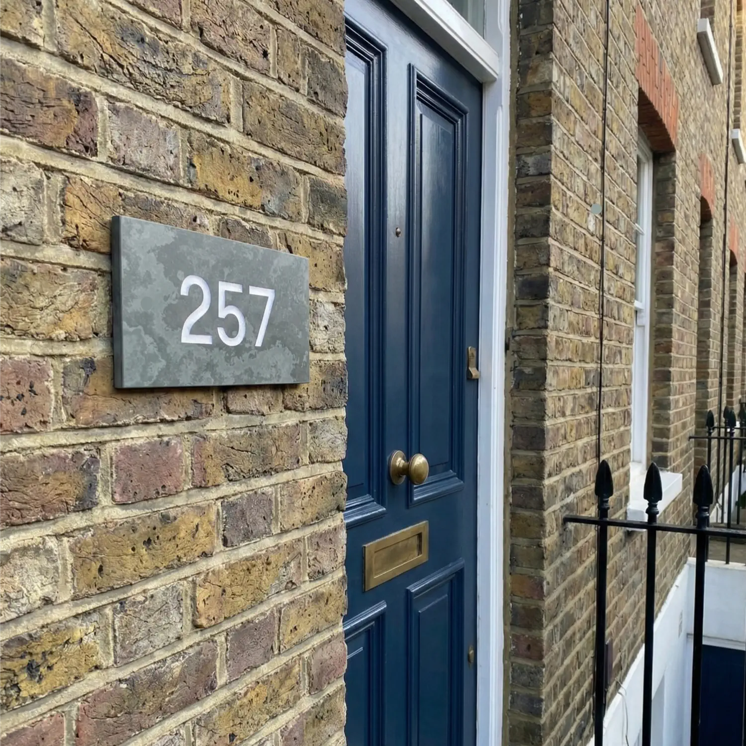 This image shows a rectangular slate sign with the number "1413" in white font mounted on a red brick wall at the corner of a building. The building features black metal fire escape stairs and balconies.