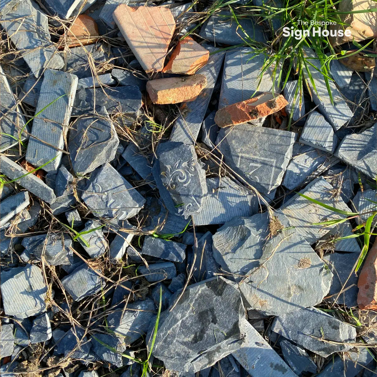 A close-up of broken slate pieces and brick fragments scattered on the ground, repurposed to fill a path. Some slate pieces feature decorative engravings, adding texture and detail amidst the grass.