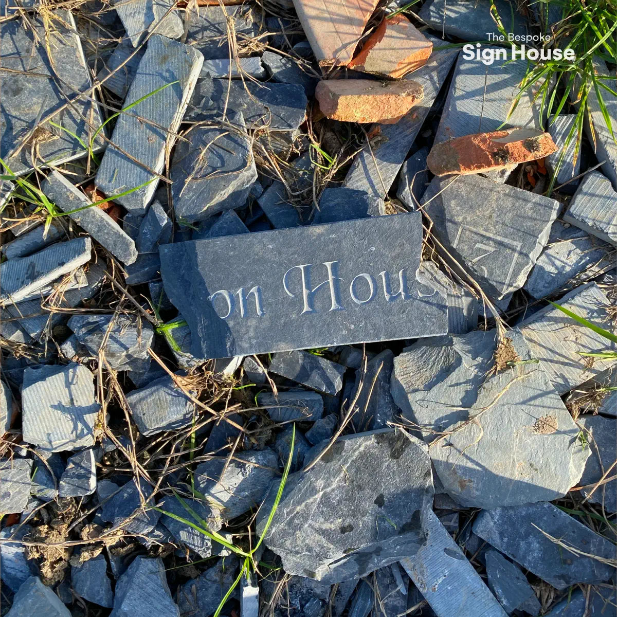 A close-up of broken slate pieces used to fill a path, with one larger piece showing partial engraving that reads “on House.” Brick fragments and bits of grass are scattered among the slate pieces.
