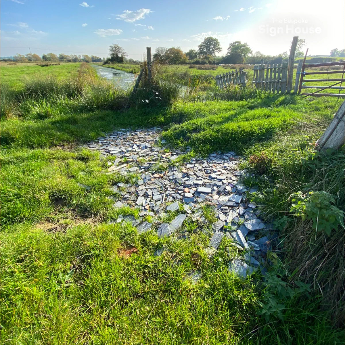 A grassy field with a path covered in broken slate pieces, used to fill a worn area. A wooden fence and a small river are visible in the background under a clear blue sky.