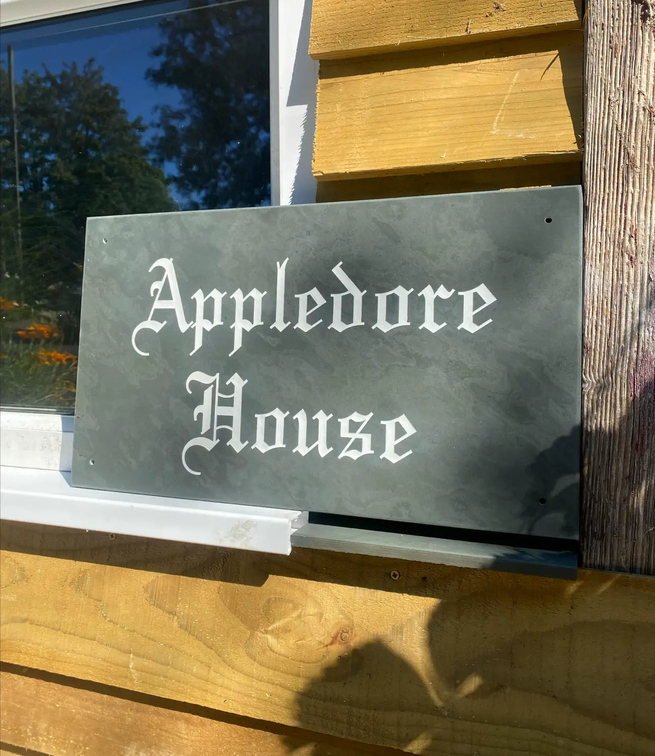 A slate house sign reading “Appledore House” in an Old English-style font, placed against a wooden-clad building with a window reflecting trees and flowers in the background.
