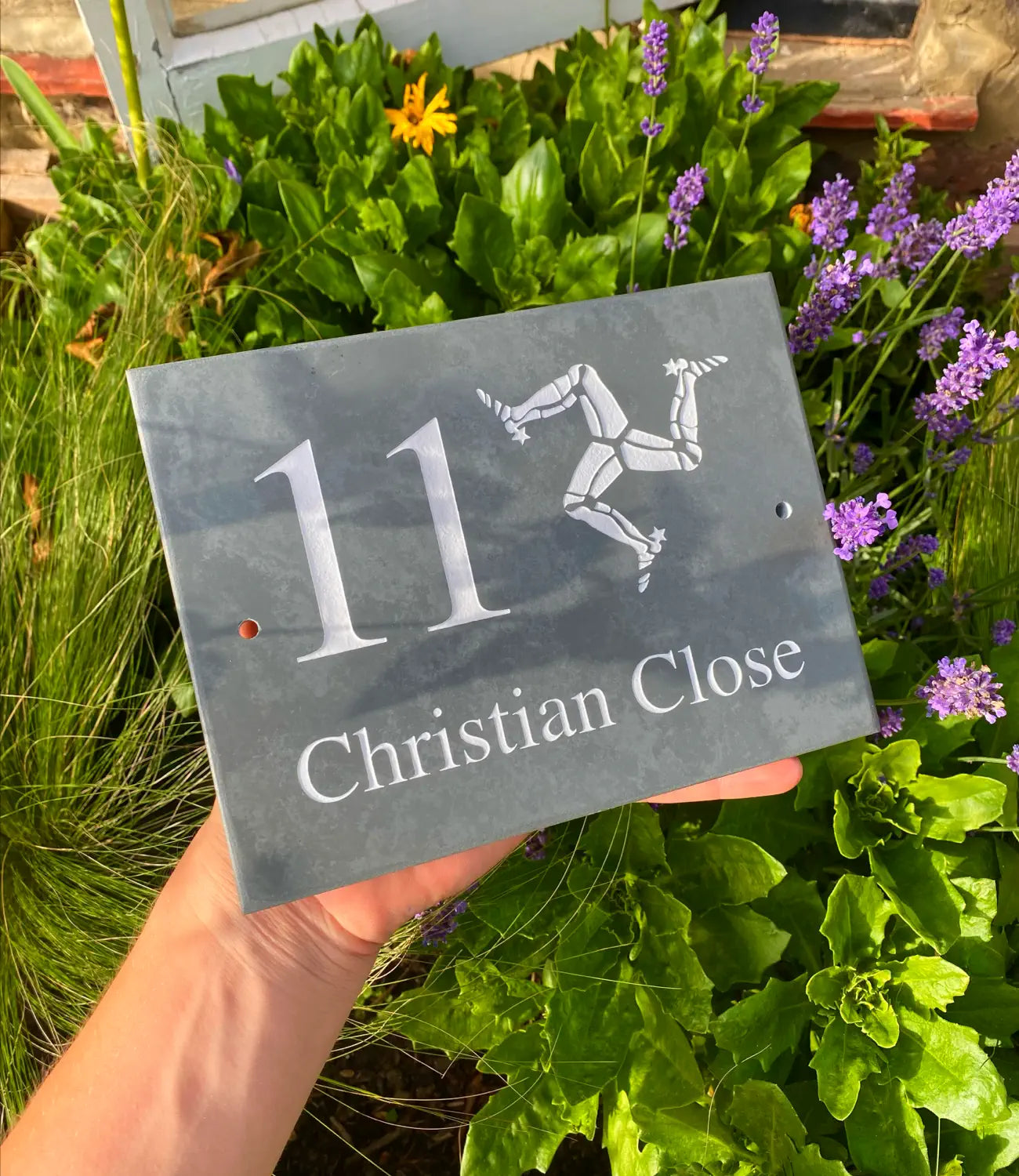 A custom slate house sign featuring the number “11” and the name “Christian Close,” engraved with the Isle of Man triskelion emblem, held outdoors against a backdrop of green plants and lavender flowers.