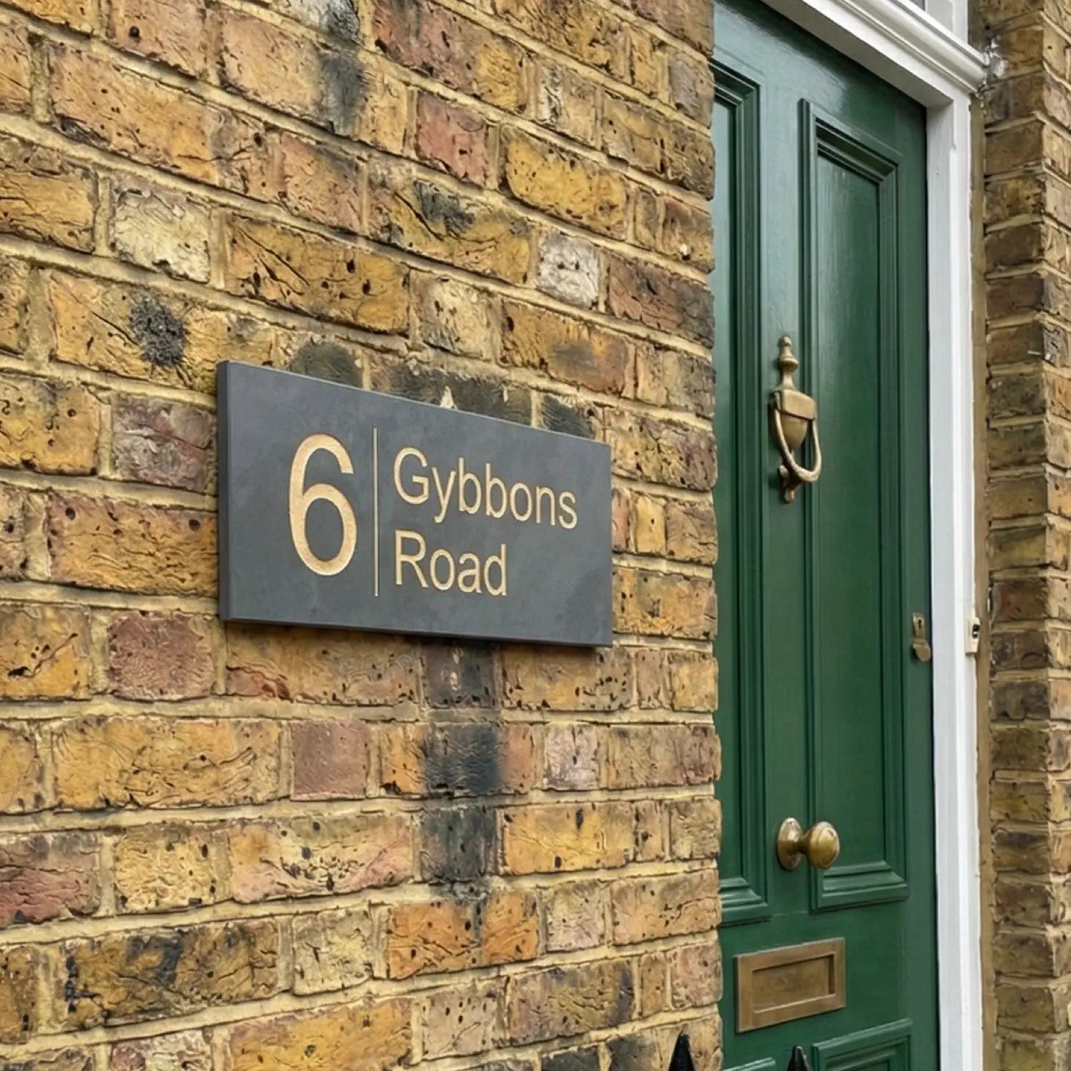 a smooth grey slate house sign with a gold number 6 and gybbons road next to it on a red brick wall
