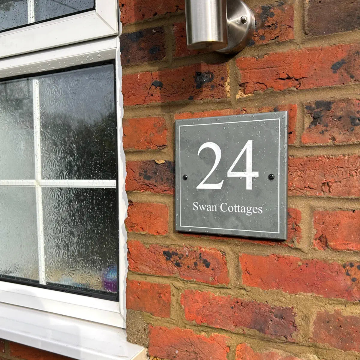 House number sign “24 Swan Cottages” mounted on a brick wall next to a frosted window and outdoor light.