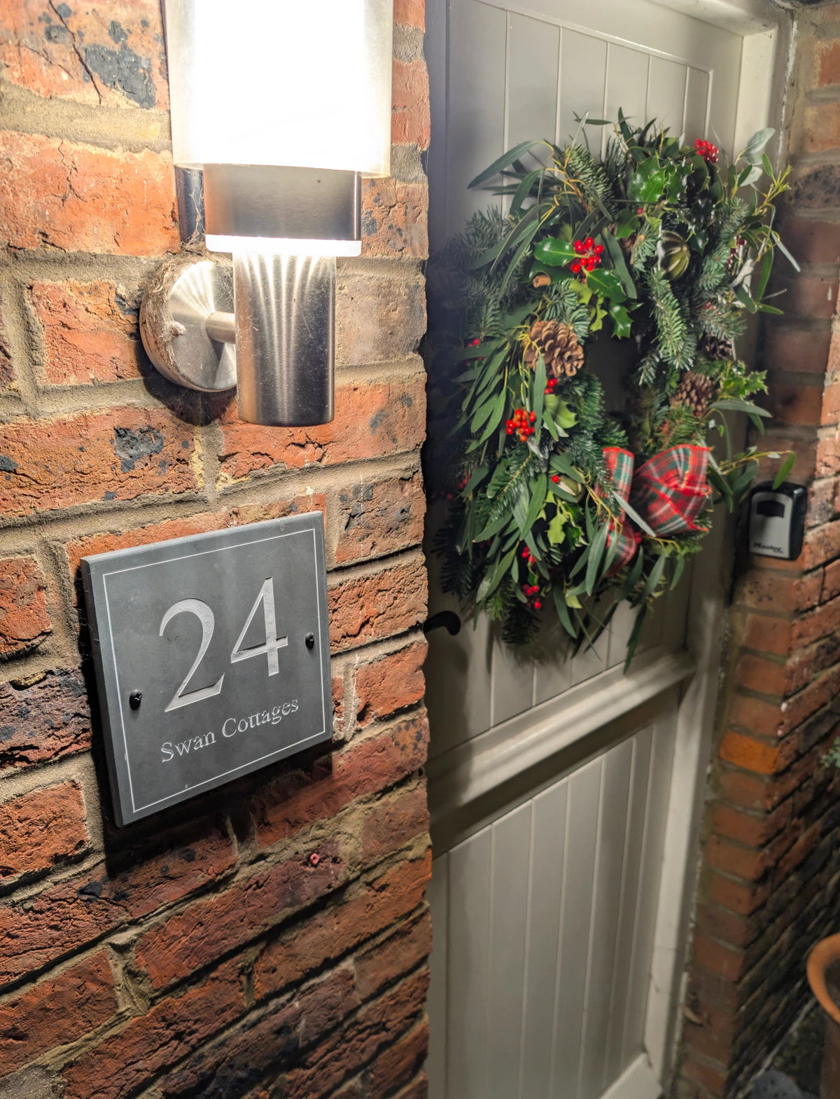 A festive green wreath with red berries and pinecones hangs on a white front door next to a red brick wall with a silver house number plaque reading '24 Swan Cottages'.