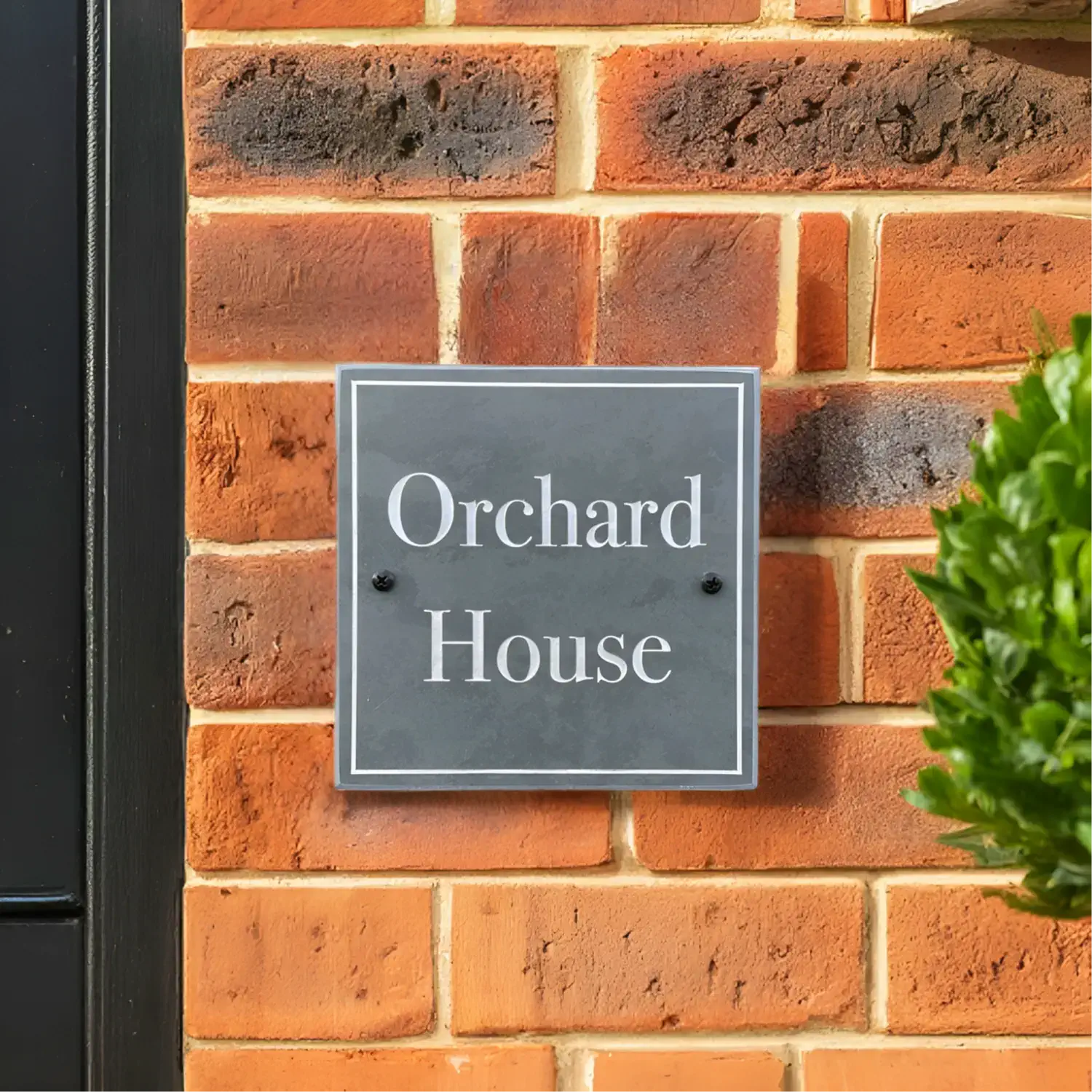 A square slate house sign with white lettering that reads “Orchard House,” mounted on a red brick wall next to a black door frame. A neatly trimmed green shrub is visible in the foreground.