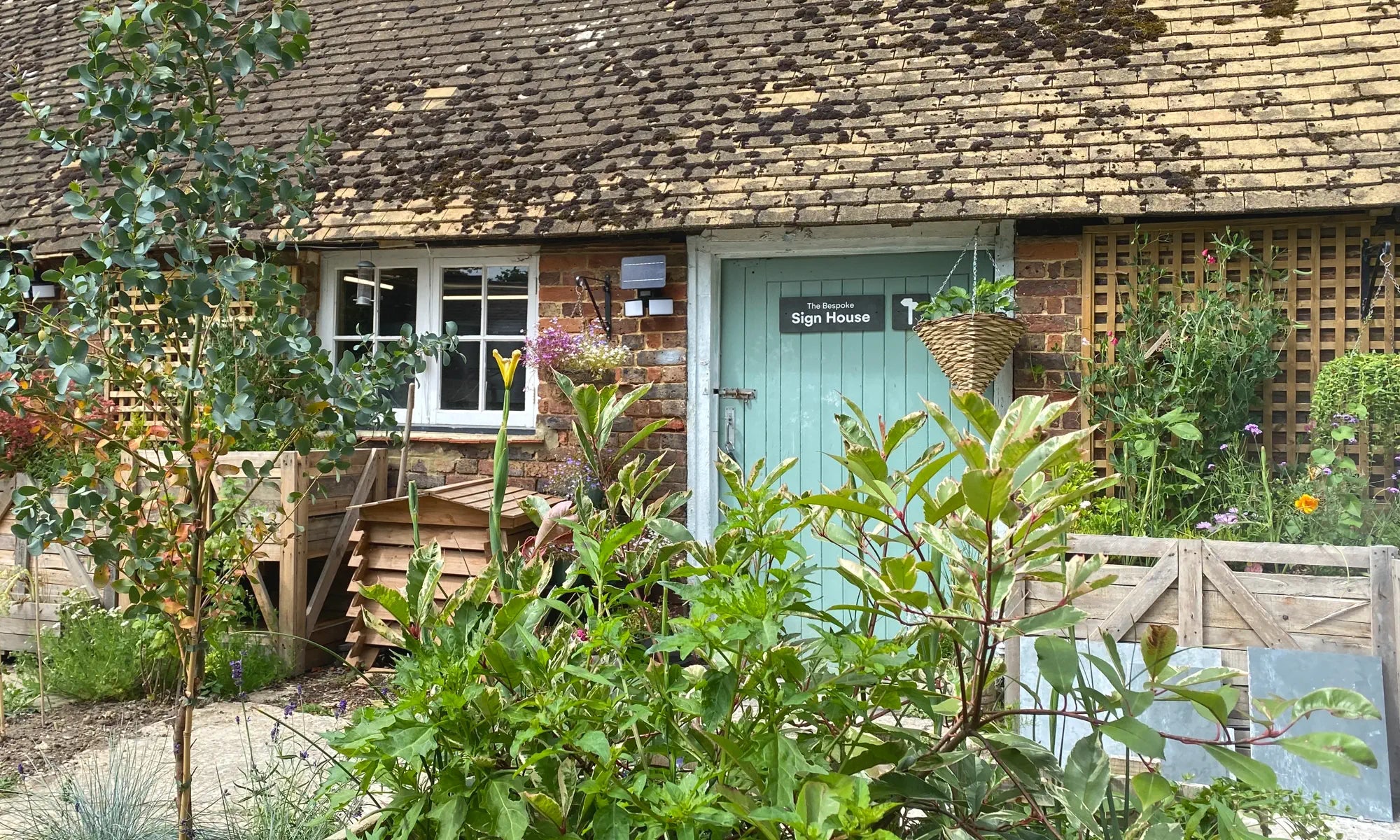 Cottage with a green door and sign, surrounded by plants and garden.