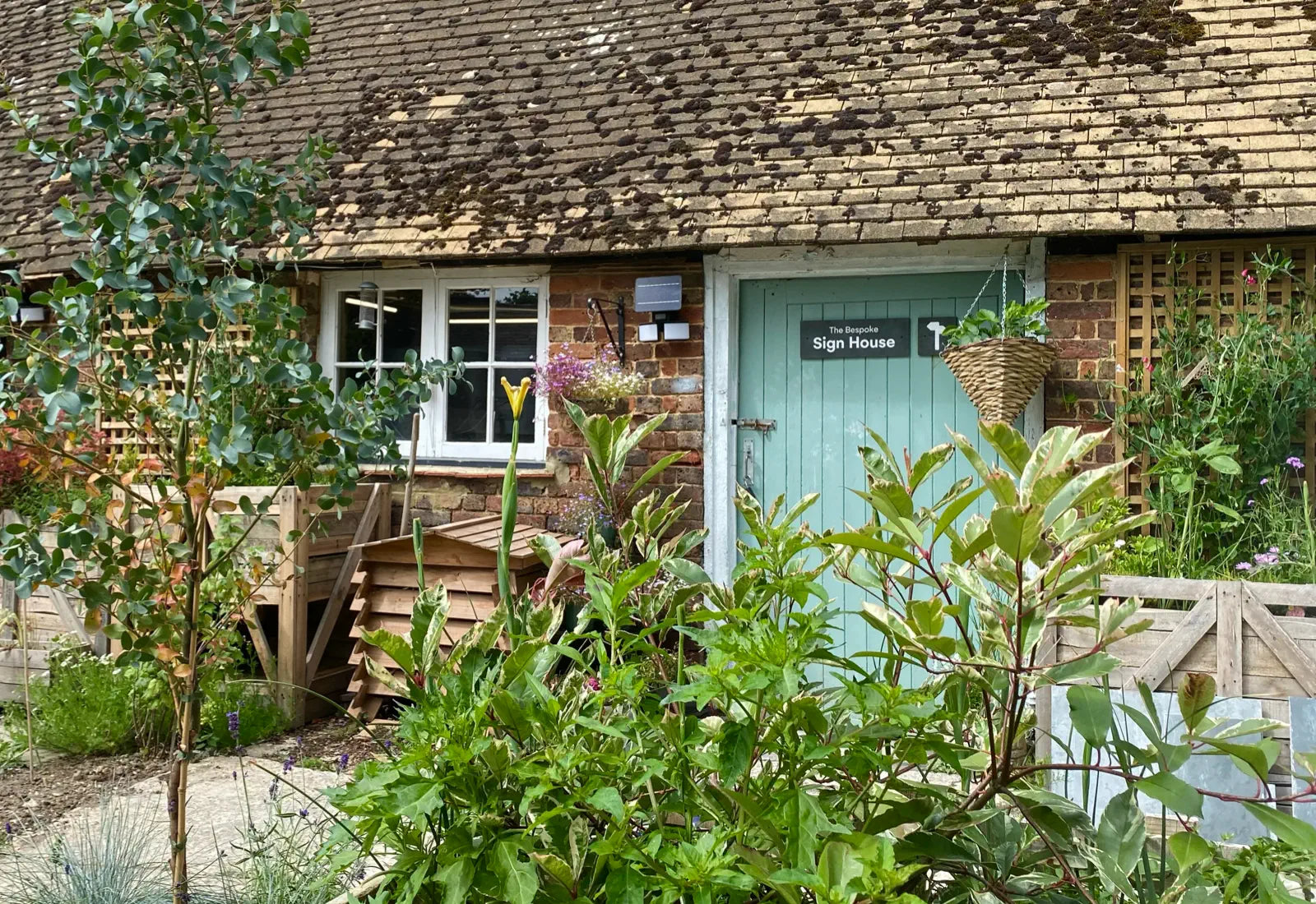 Cottage with a green door and sign, surrounded by garden plants and flowers.