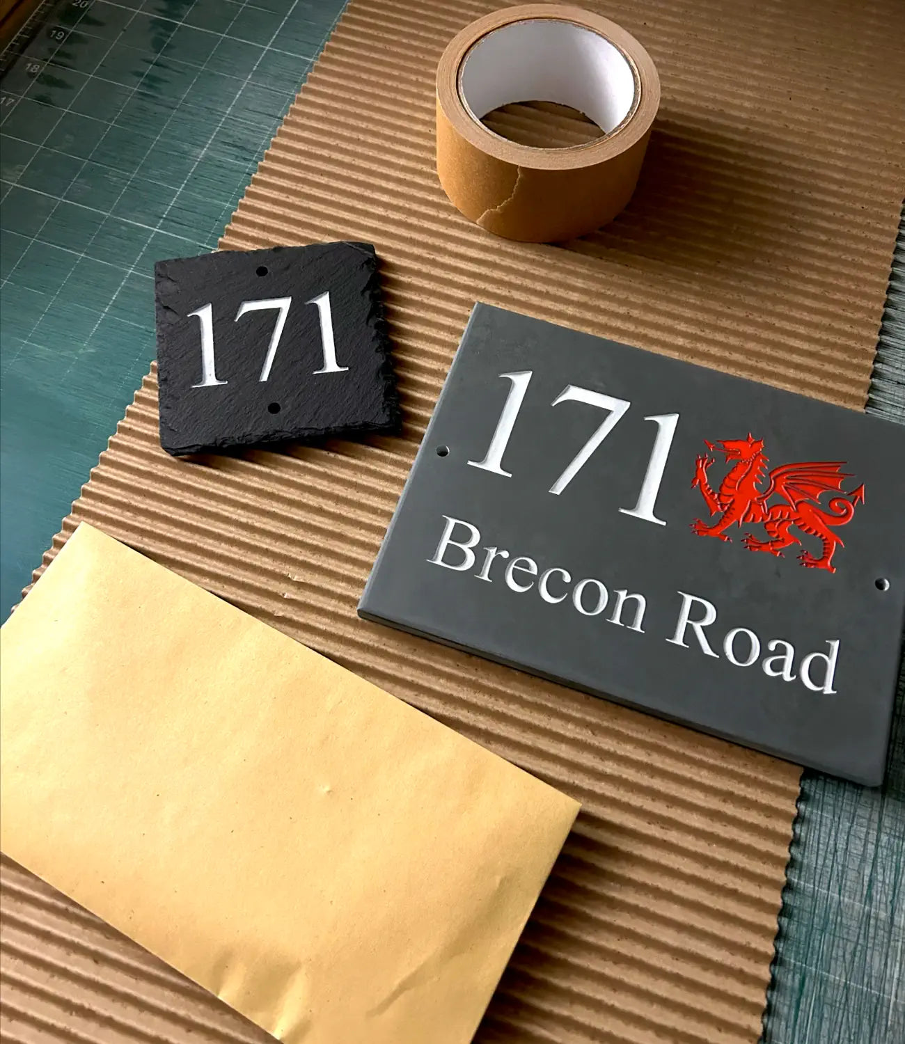 Two custom slate house signs, one featuring the number “171” and the other engraved with “171 Brecon Road” alongside a red Welsh dragon emblem. Displayed on a packaging surface with brown paper, cardboard, and tape, ready for wrapping and shipping.