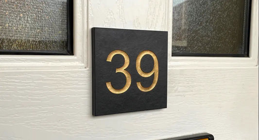 a small square dark grey slate sign glued onto a white upvc door. The number is 39 which is painted gold