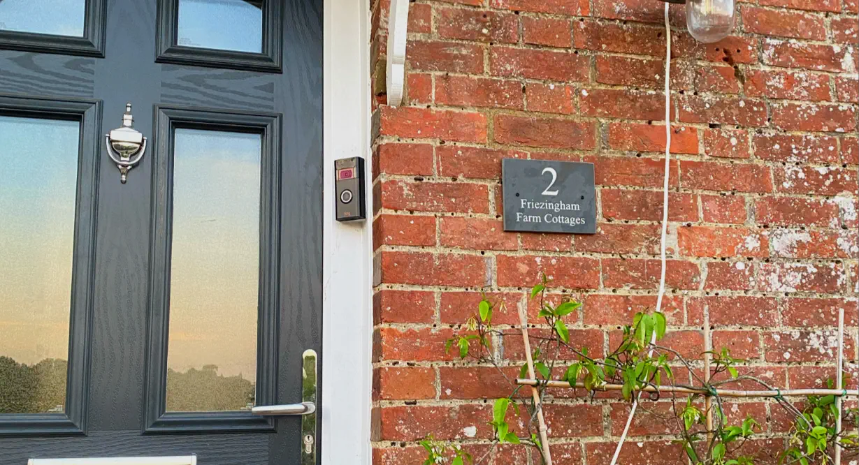 The entrance to “2 Friezingham Farm Cottages” features a dark gray door with a silver knocker, a modern doorbell, and a house plaque on a red brick wall. A climbing plant on a trellis and a simple outdoor light complete the scene.