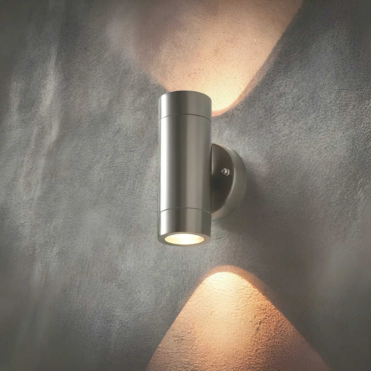 Sleek stainless steel outdoor wall light with up-and-down illumination, casting warm light against a textured grey wall.