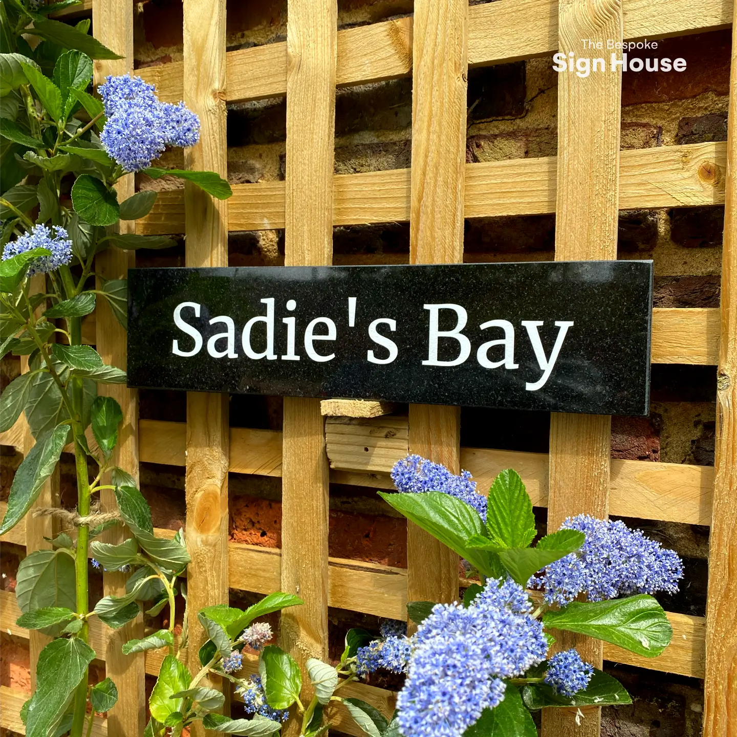 Granite house sign reading ‘Sadie’s Bay’ mounted on a wooden lattice with blooming blue flowers and green leaves surrounding the sign. The sign is set against a rustic brick wall, adding a natural and elegant touch to the garden setting.