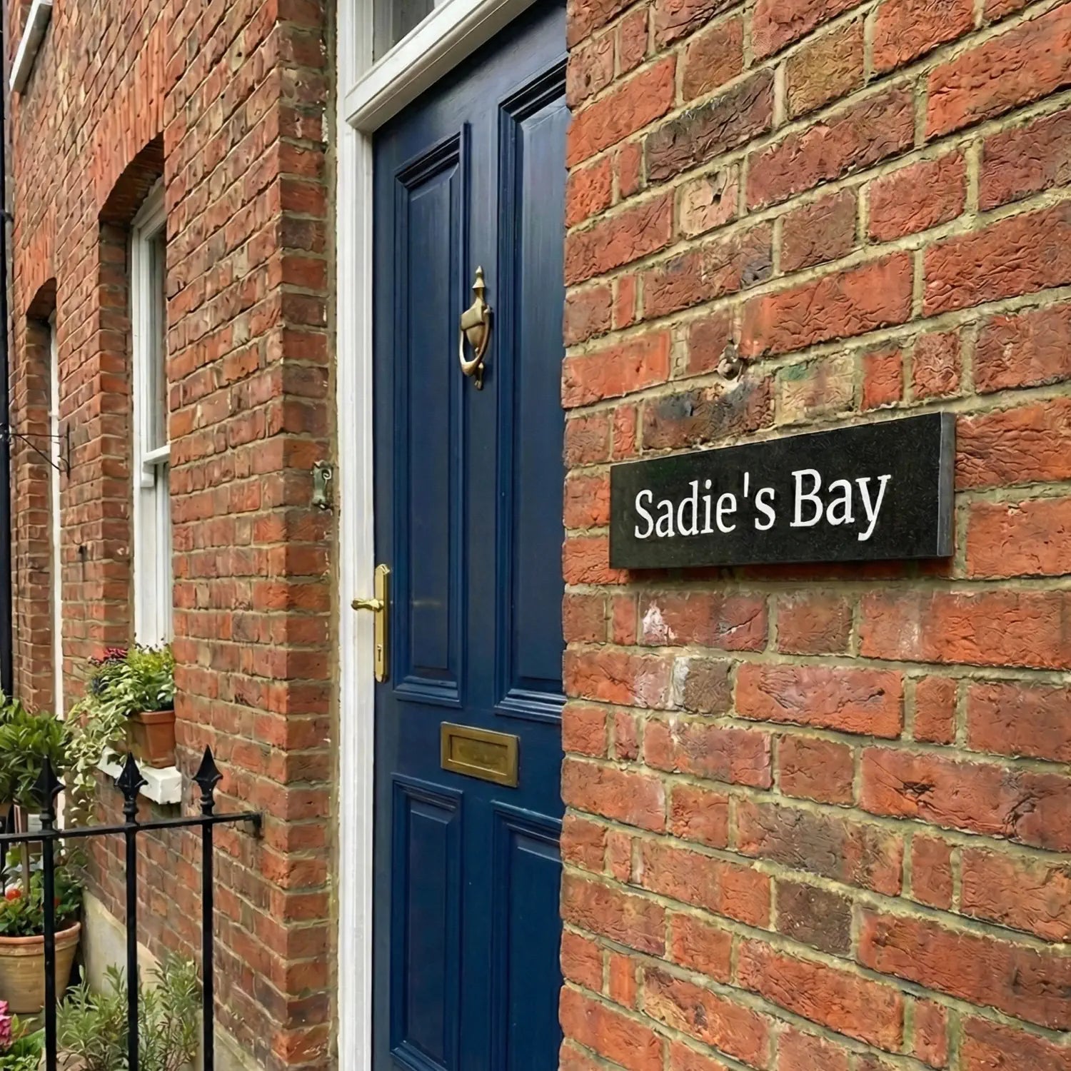 A black granite house sign with white text reading 'Sadie's Bay', mounted on a wooden lattice with purple flowers in the foreground. 