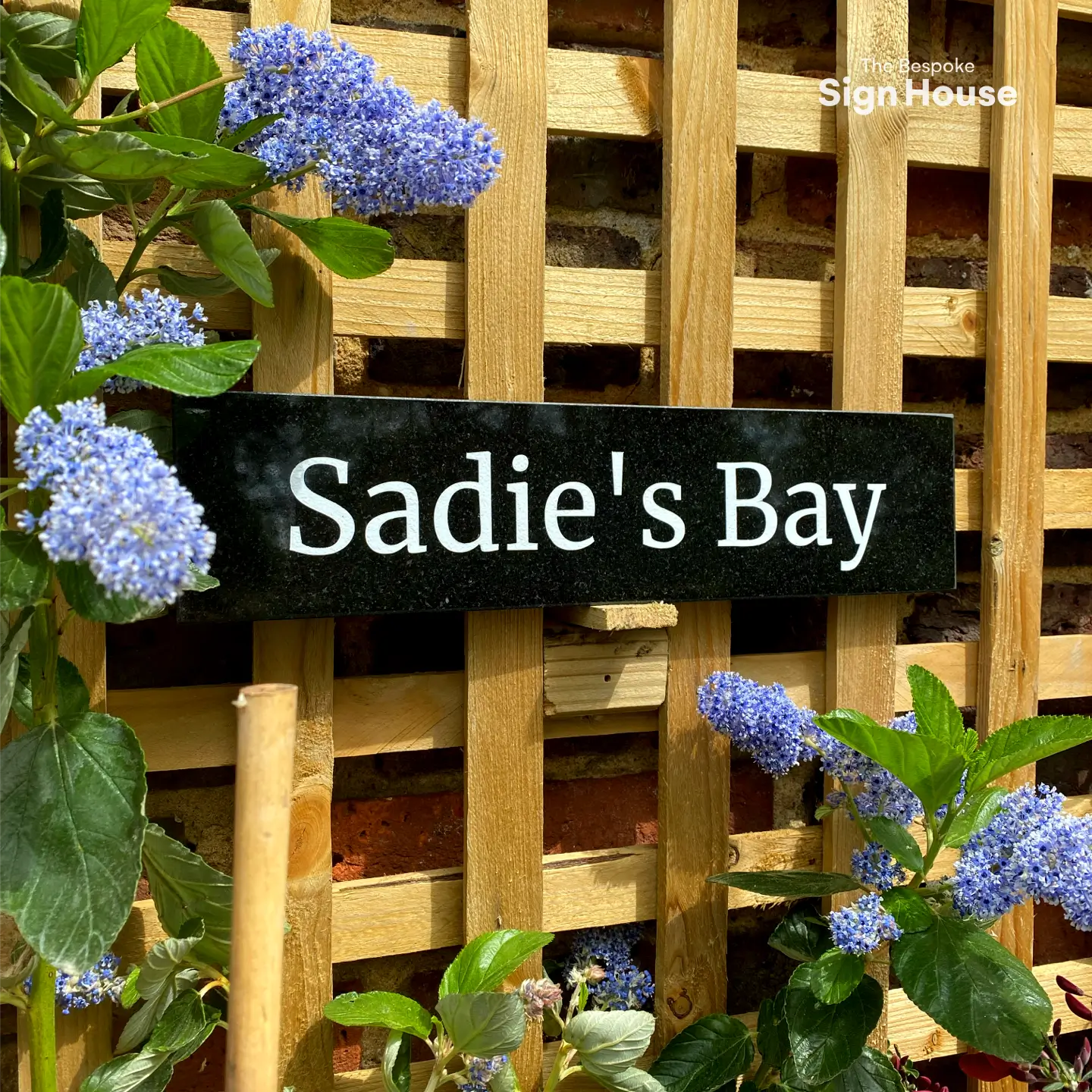 Granite house sign with the text ‘Sadie’s Bay’ mounted on a wooden lattice, surrounded by vibrant green leaves and clusters of blooming blue flowers. The sign is placed against a brick wall, creating a charming and natural garden atmosphere.