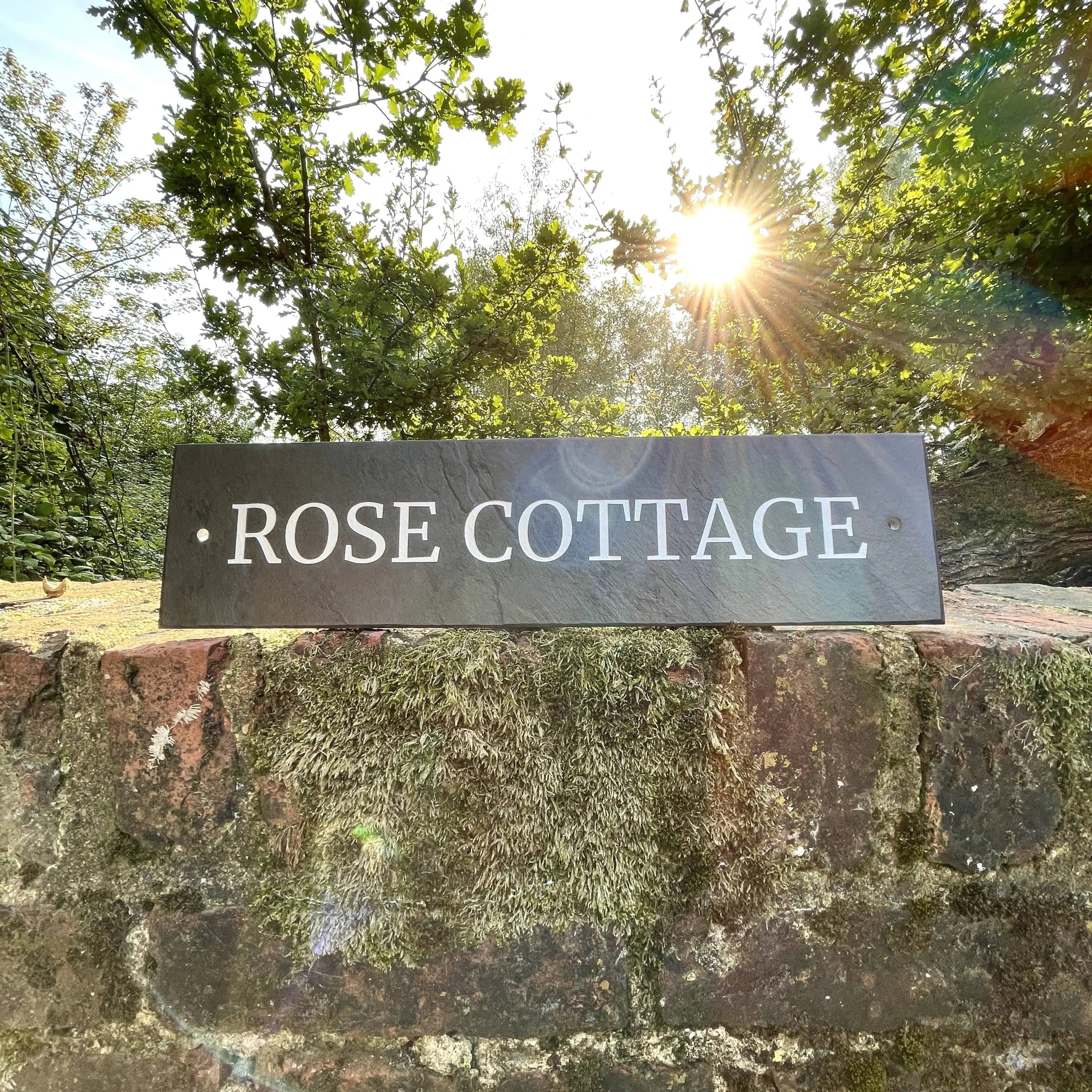 Sign reading 'Rose Cottage' on a stone wall with trees in the background