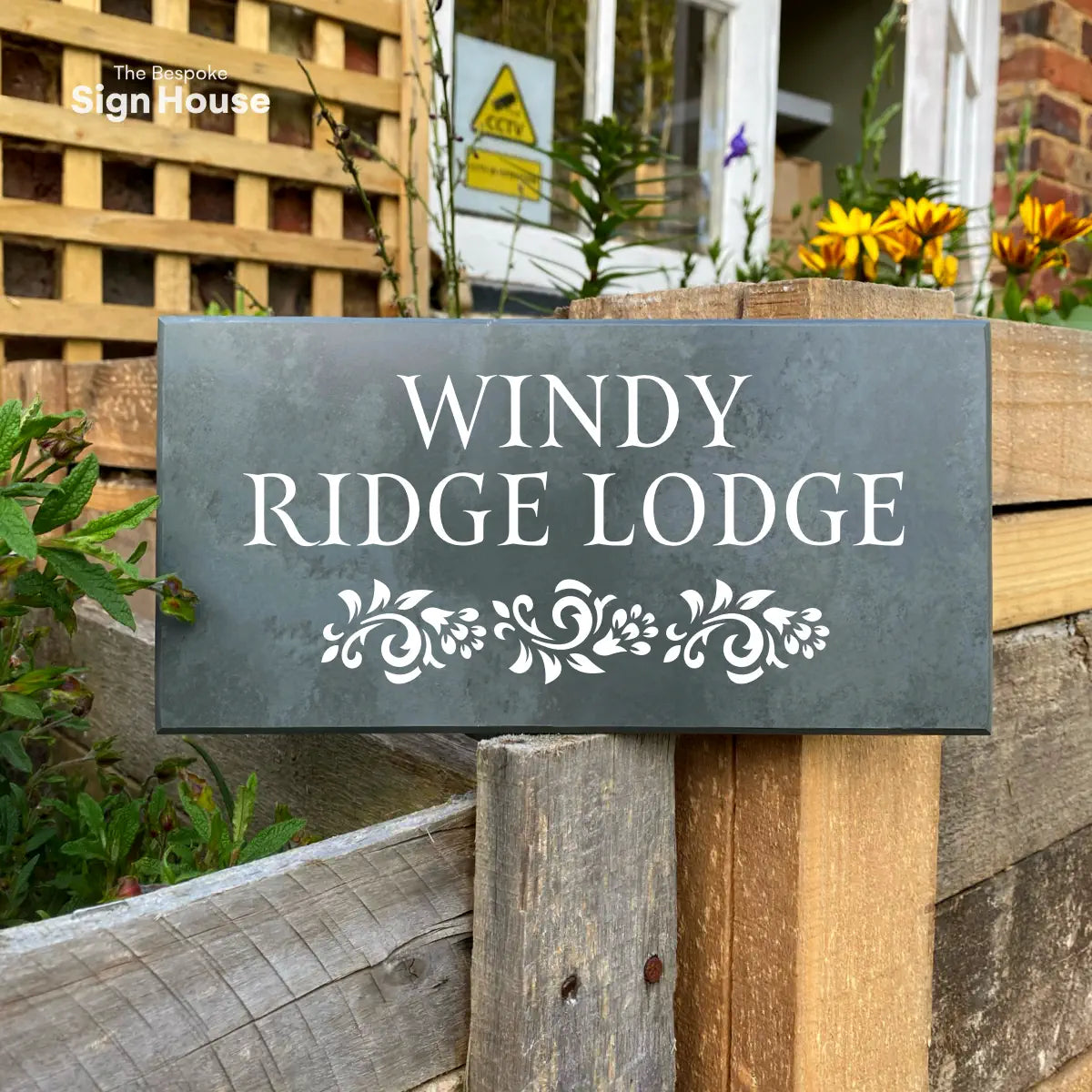 A slate house sign positioned on a wooden frame. The sign featured some floral detailing at the bottom in white. Above has the customers address engraved in white also. The sign reads Windy Ridge Lodge