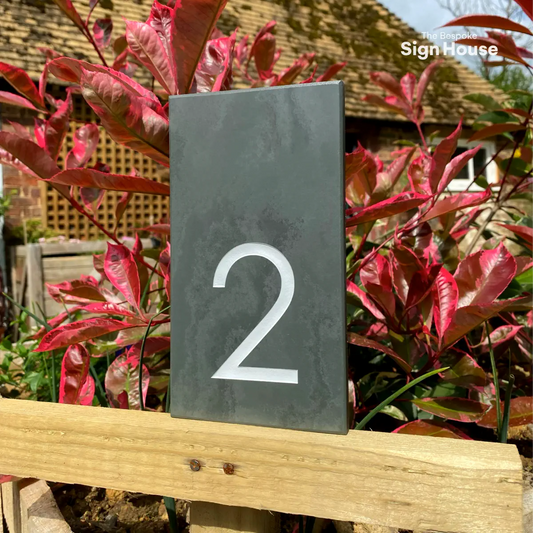 a grey slate house number sign with the number 2 engraved and painted white. The sign is against a backdrop of red leaves