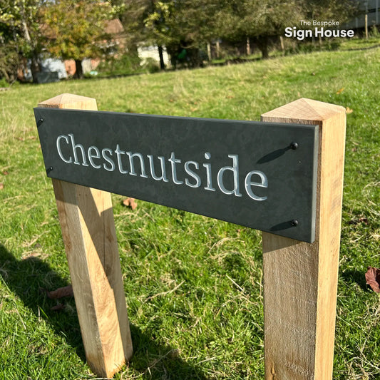 The image shows a slate house sign mounted onto sturdy oak posts with pyramid tops. The sign reads “Chestnutside” in clear white lettering, and the oak posts are set into a grassy field. The overall appearance is stylish and rustic, showcasing the natural wood and elegant slate combination. The Bespoke Sign House logo is visible in the top right corner.