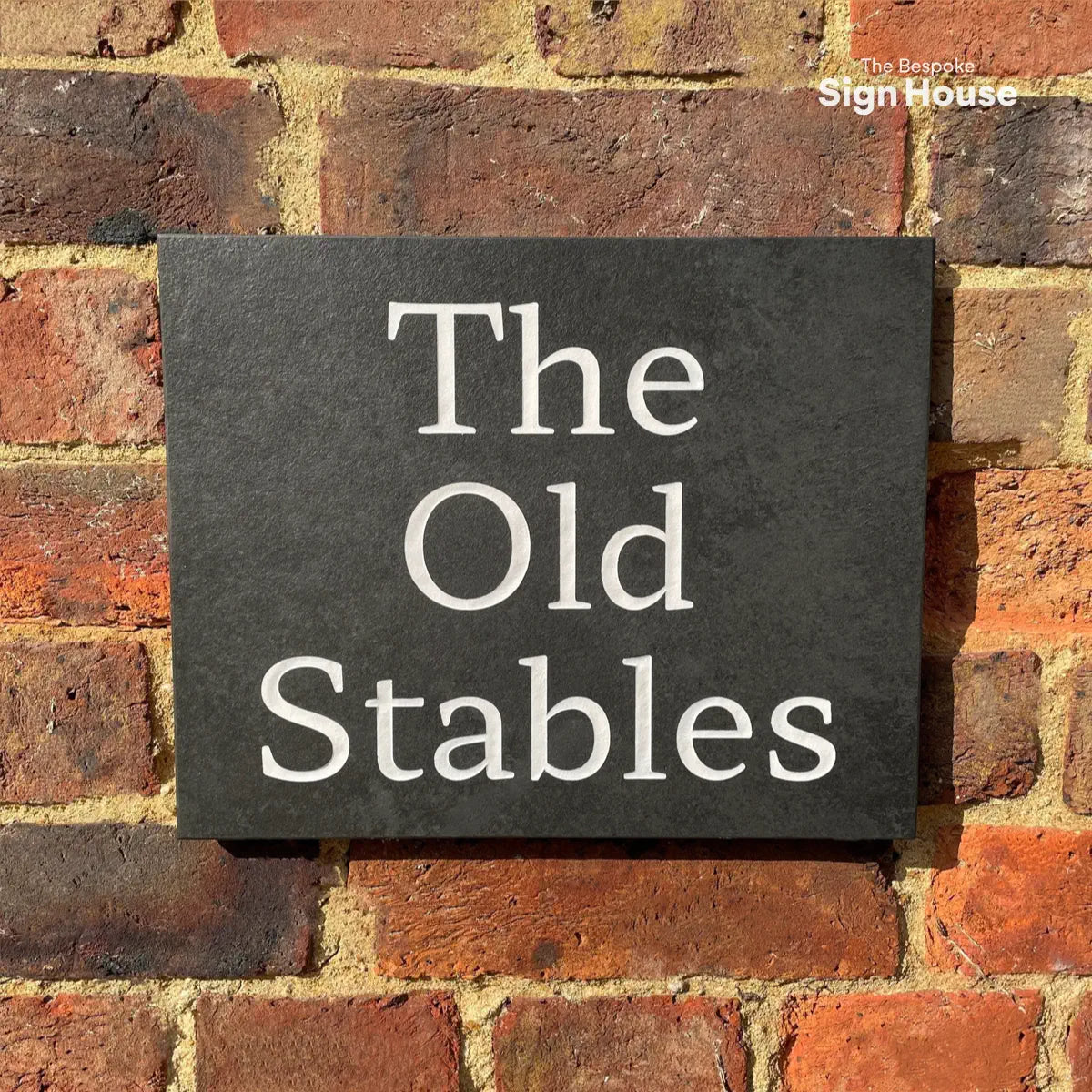 A dark slate house sign with white lettering that reads “The Old Stables,” mounted on a rustic red brick wall.