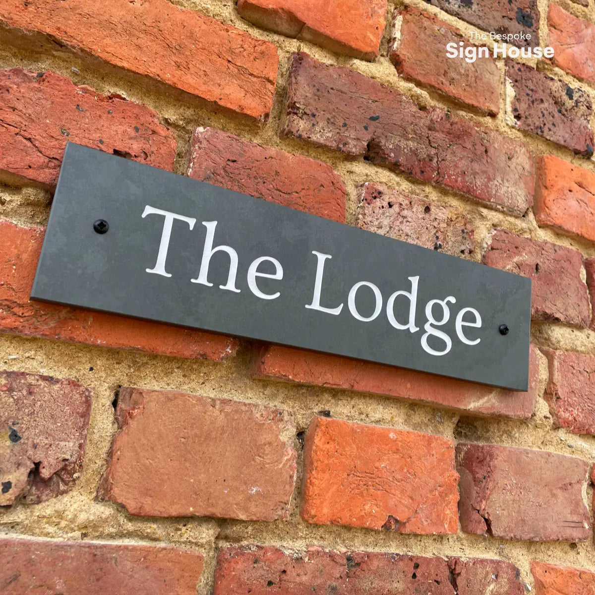 A rectangular dark slate house sign with white text that reads “The Lodge,” mounted on a textured red brick wall.