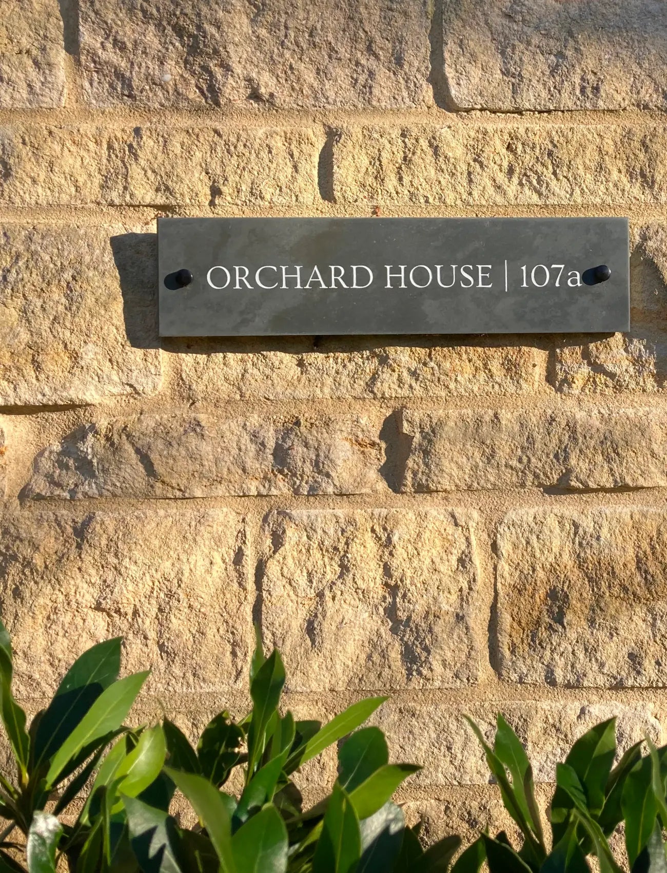 Slate house sign on a stone wall with 'Orchard House | 107a' text.